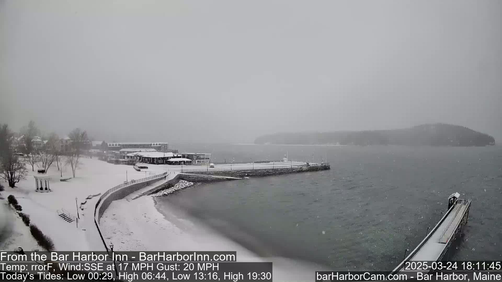 A snow-covered waterfront scene shows a harbor with a long pier extending into the water, buildings in the background, and a lightly snowing sky.