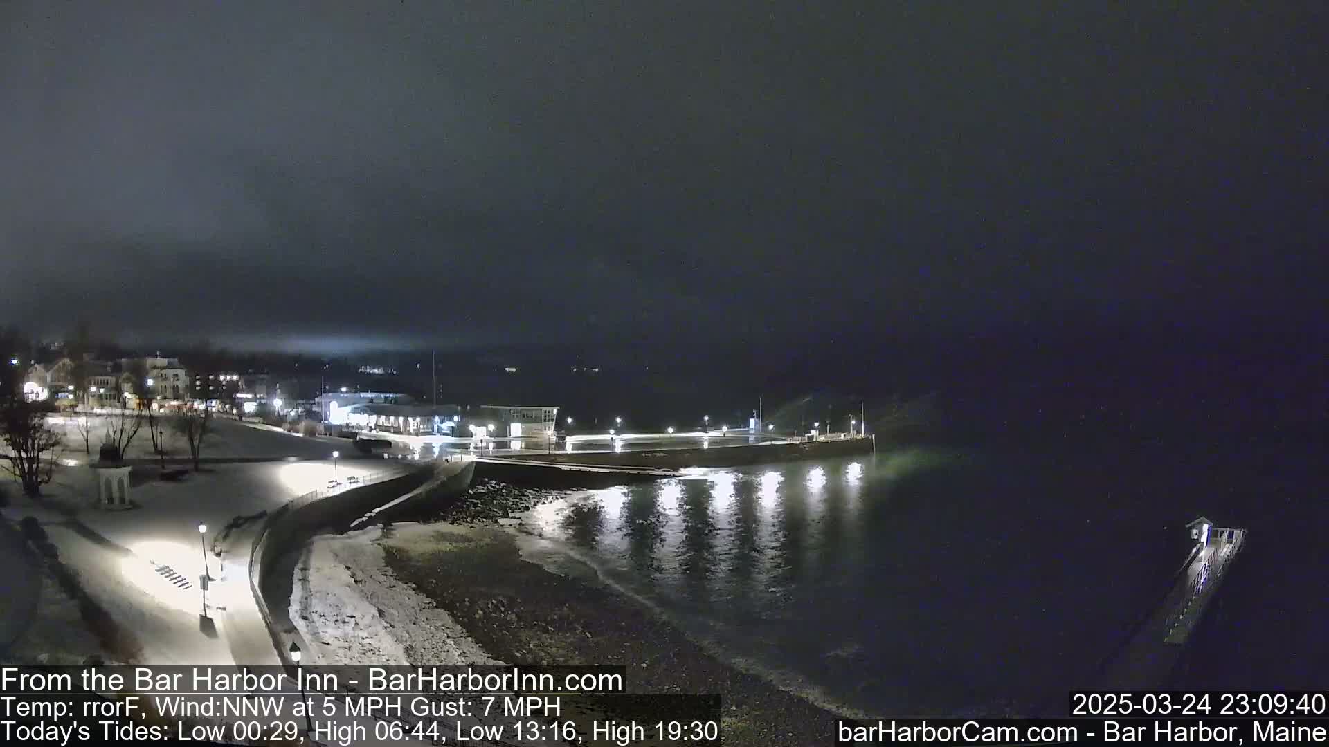 A nighttime coastal scene shows a snow-covered shoreline with lights illuminating buildings and a pier extending into dark, gently lapping water under a cloudy sky.