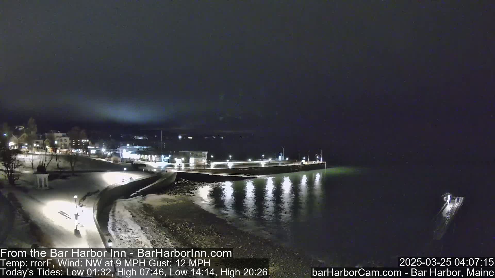 A nighttime view of a snow-covered harbor town with a pier extending into calm dark water under a dark, cloudy sky.