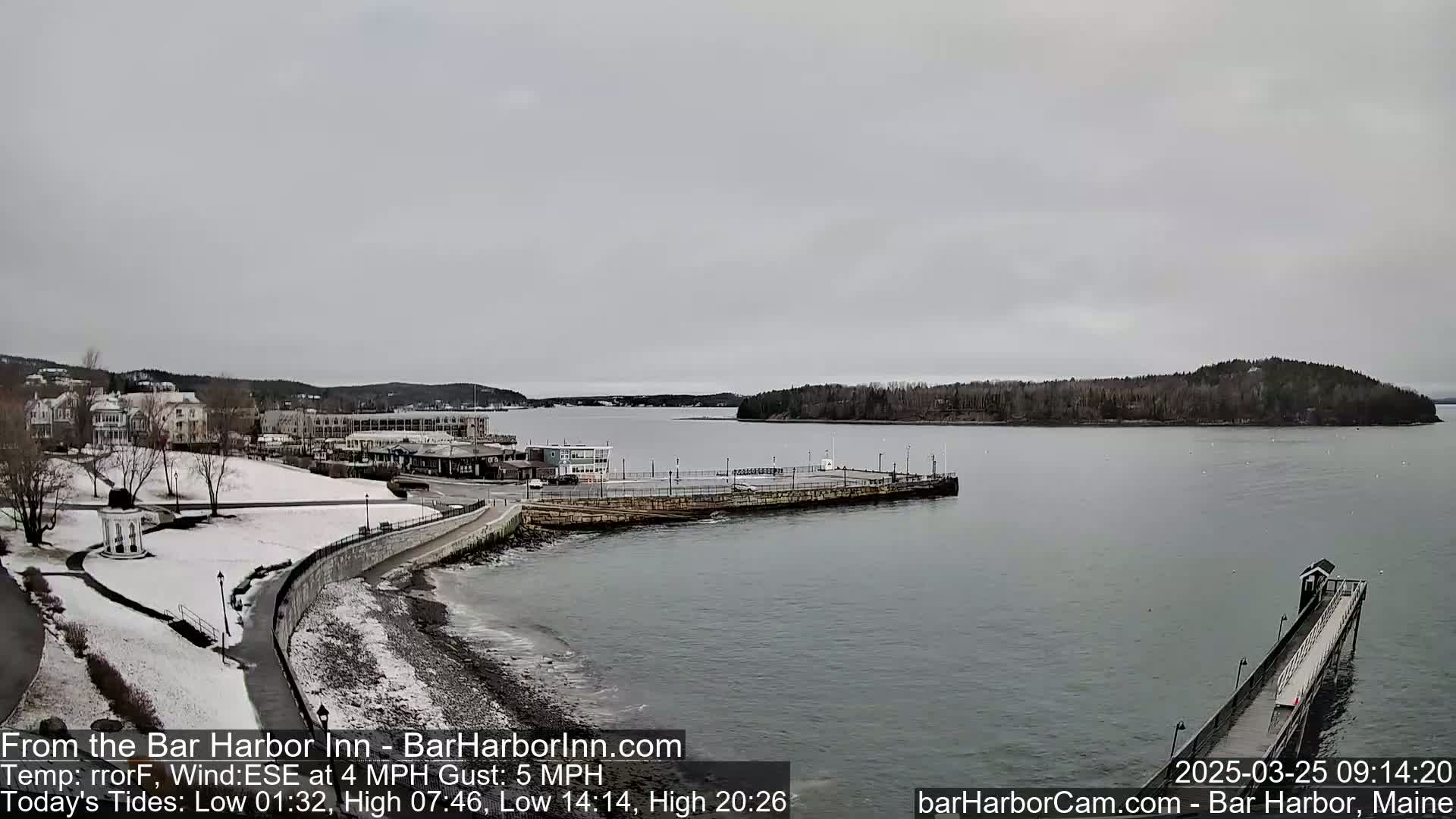 A snow-dusted coastal town is seen under an overcast sky, with a calm body of water featuring a long pier extending into the bay.