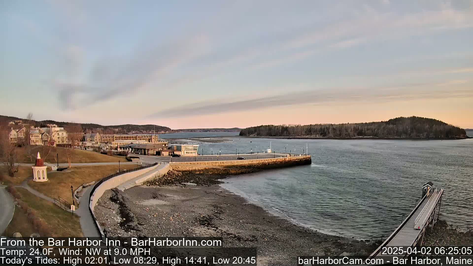 A coastal scene at sunrise shows a calm harbor with a long pier extending into the water, buildings in the background, and a small gazebo on the shore.