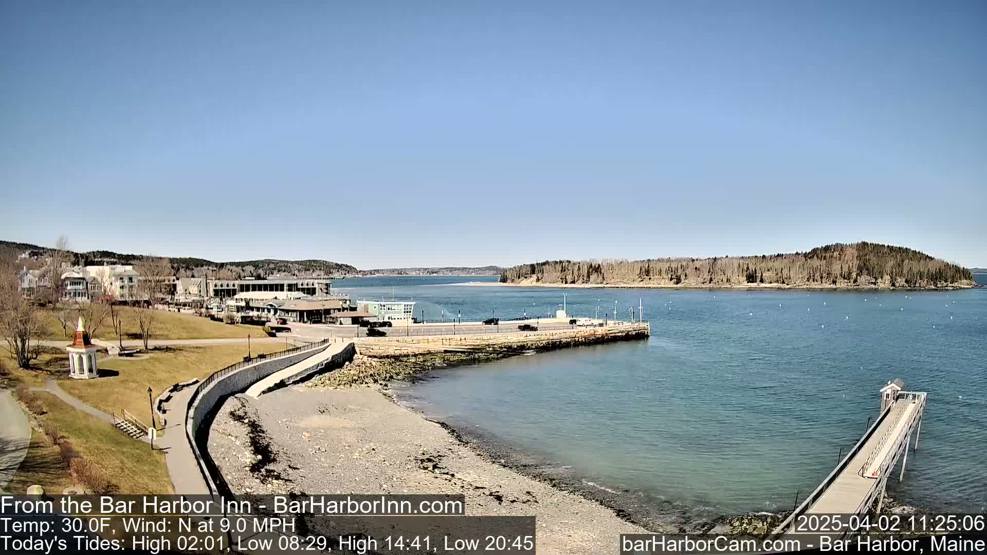 A clear, sunny day shows a rocky shoreline with a calm harbor, a small town in the background, and a long pier extending into the water.