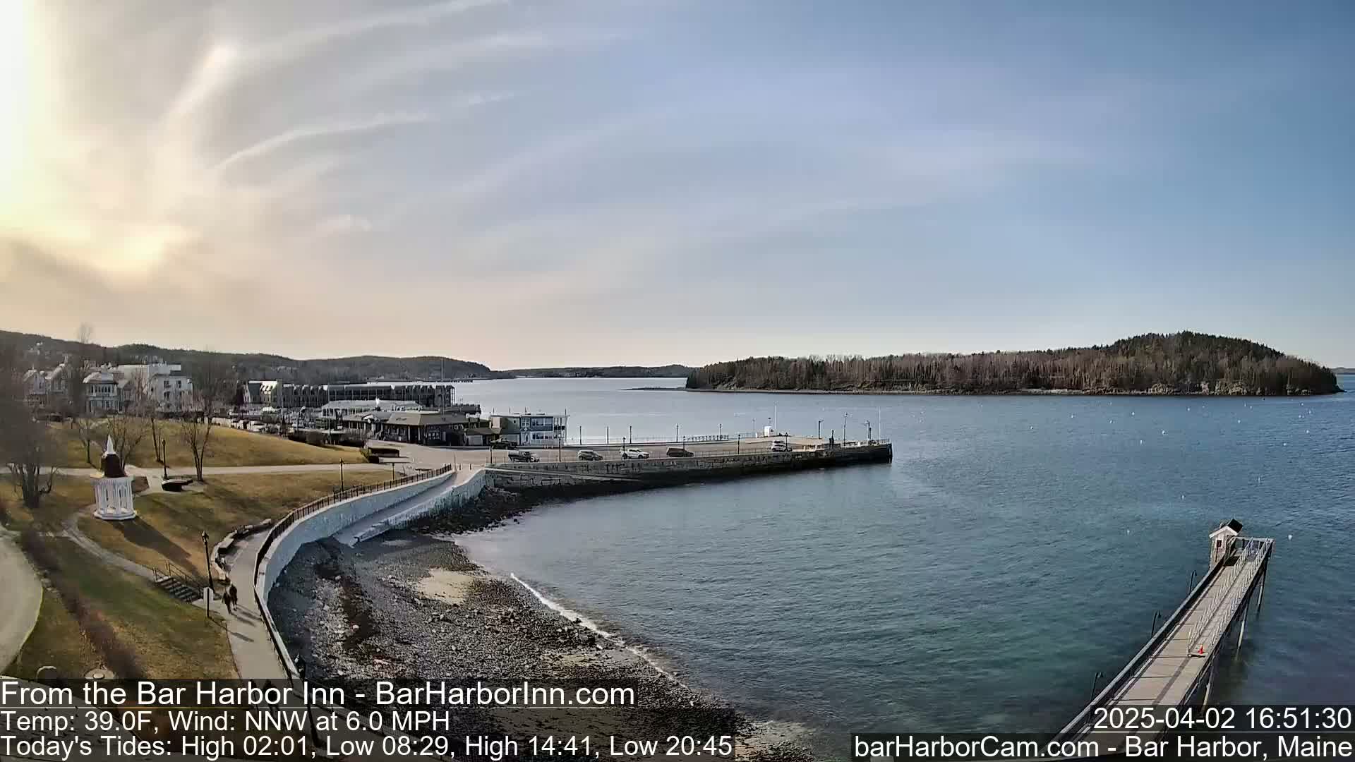 A partly sunny day shows a coastal town with a harbor, a pier, and a small island in the distance.