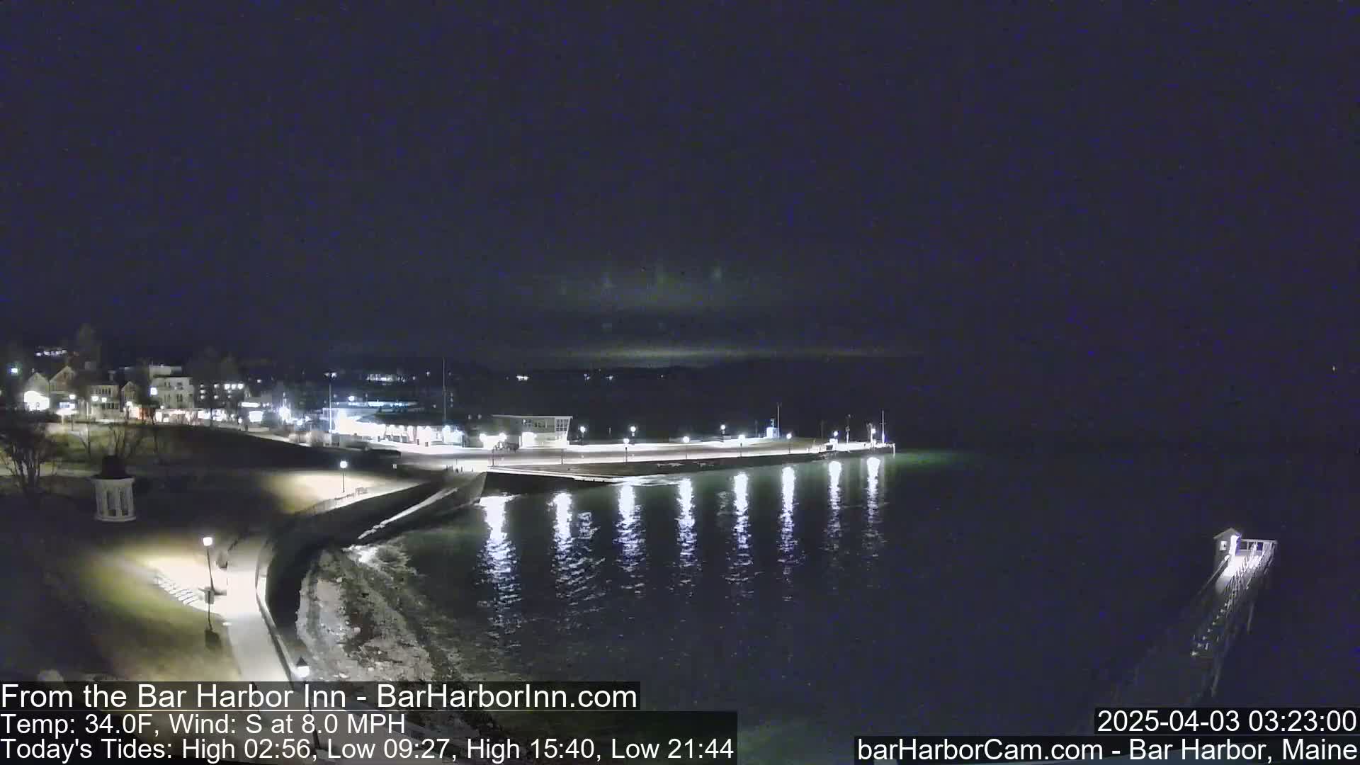 A nighttime coastal scene shows a town's lit shoreline, a calm dark-water harbor with reflections of lights, and a pier extending into the water under a clear, dark sky.