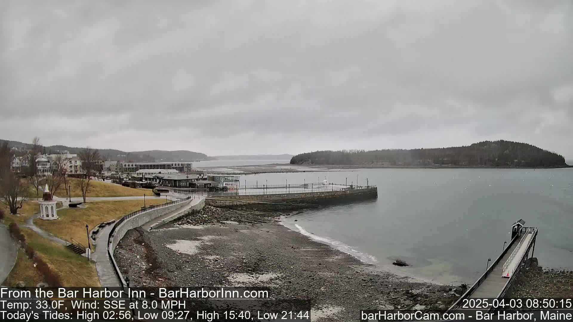 A coastal town scene under an overcast sky shows a rocky shore, calm water, a long pier, and buildings in the distance.