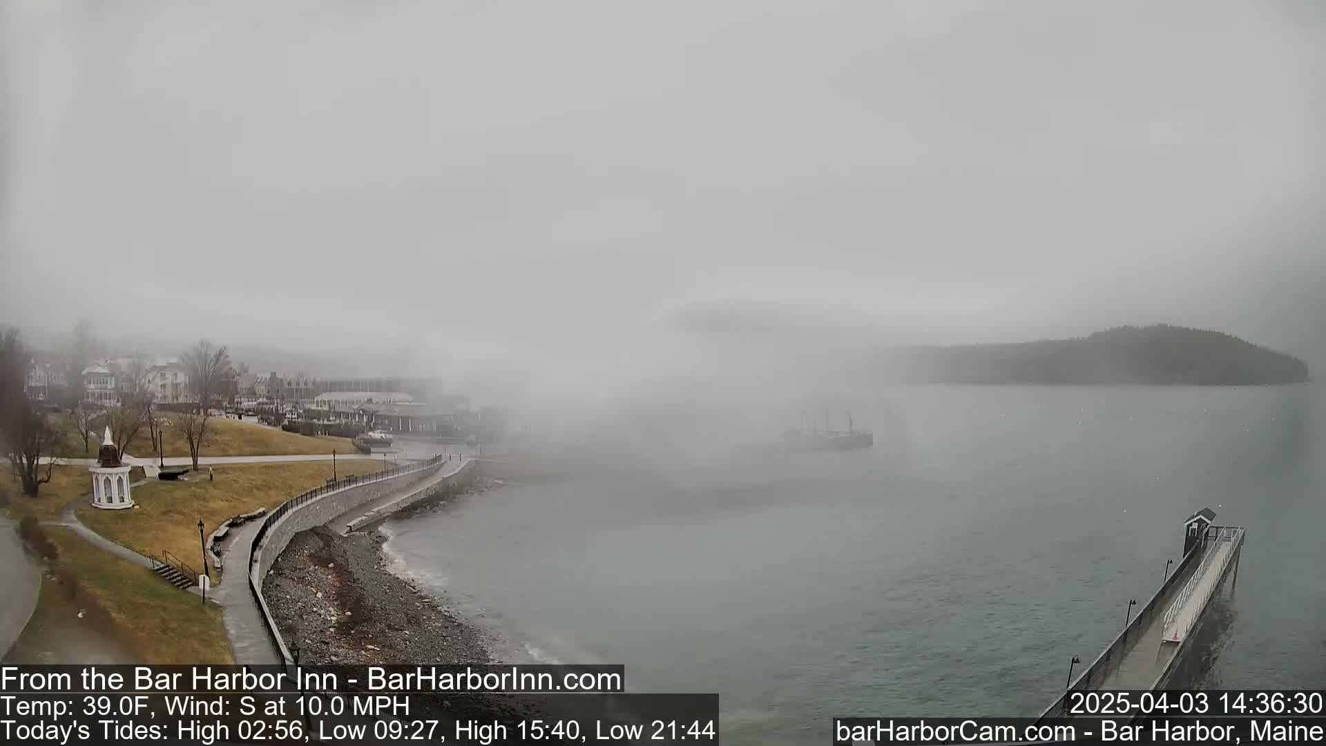 A foggy coastal scene shows a town, a shoreline with a walkway, a small island in the distance, and a long pier extending into the water.