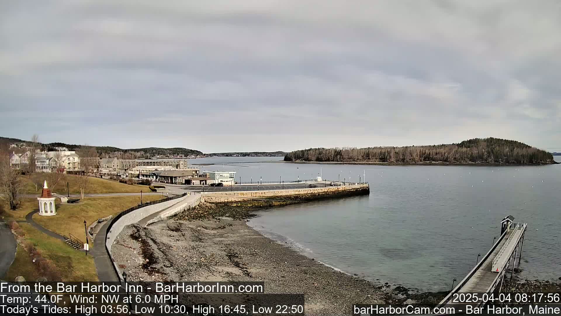 A coastal town with buildings and a grassy area overlooks a calm harbor with a long pier extending into the water under an overcast sky.