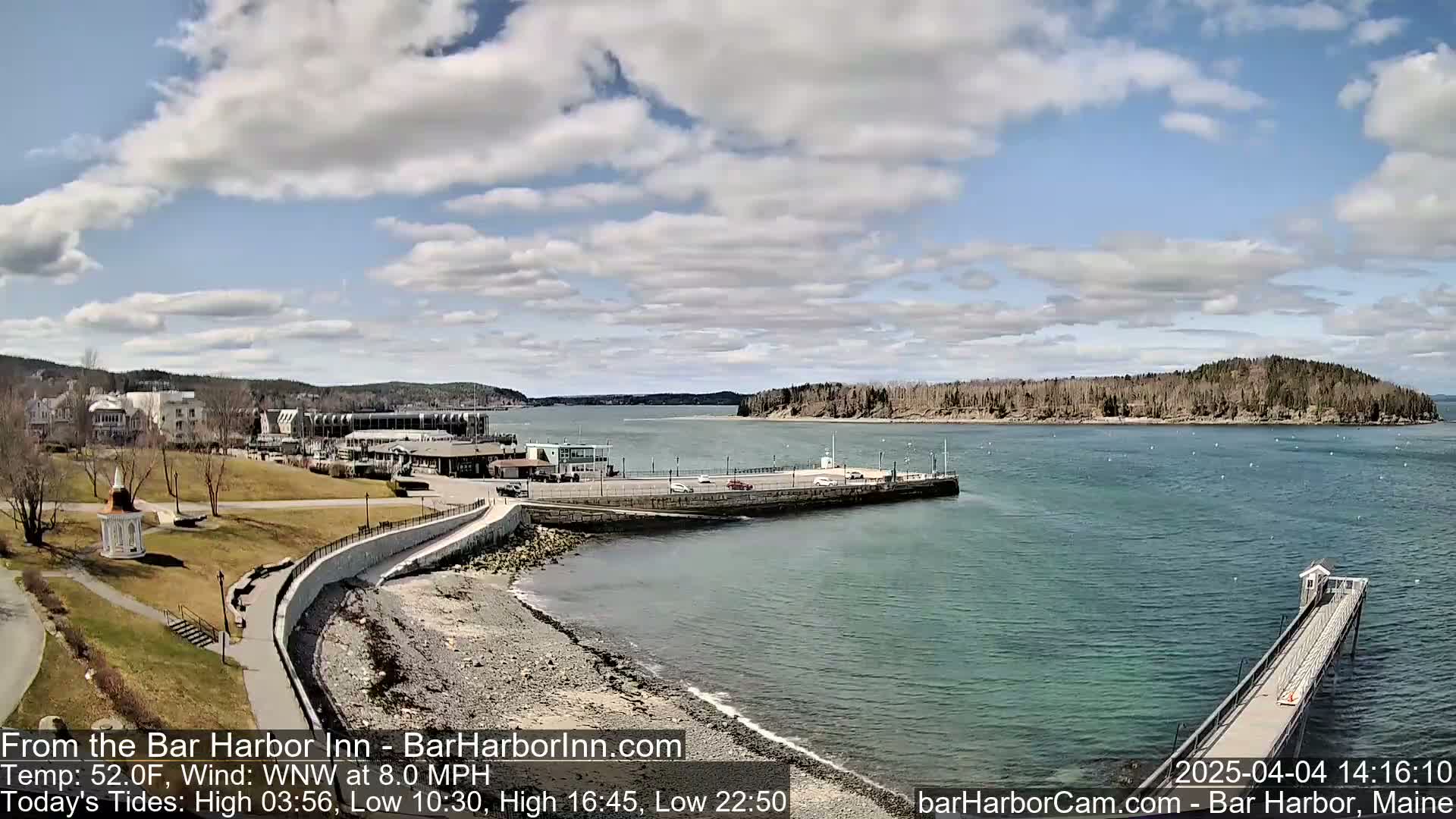 A partly cloudy day shows a rocky shoreline with a town and harbor in the background, a long pier extending into the water, and a small island visible in the distance.