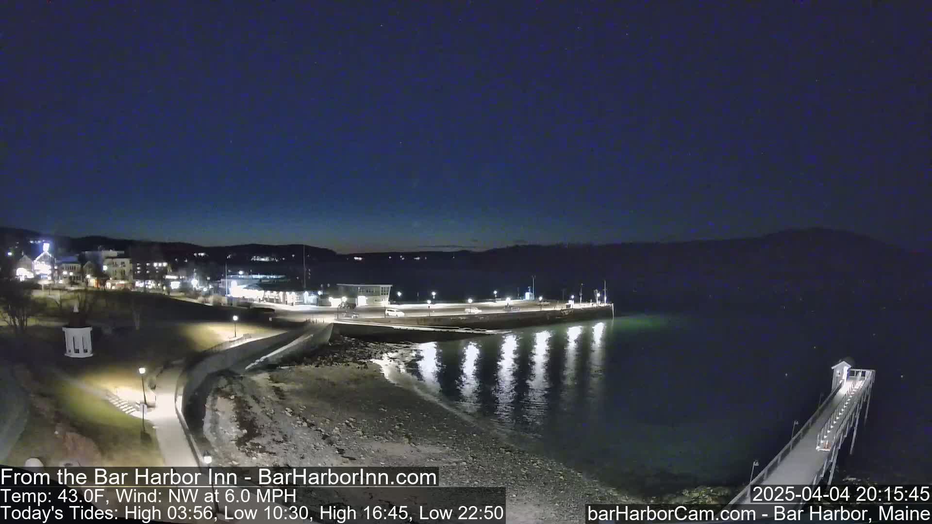 A nighttime coastal scene shows a town illuminated along a calm, dark body of water, with a long pier extending into the water.