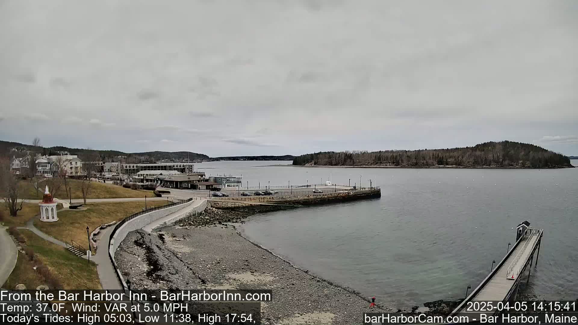 A waterfront town with a stone walkway, a small gazebo, and a long pier extending into a calm, gray-sky day.
