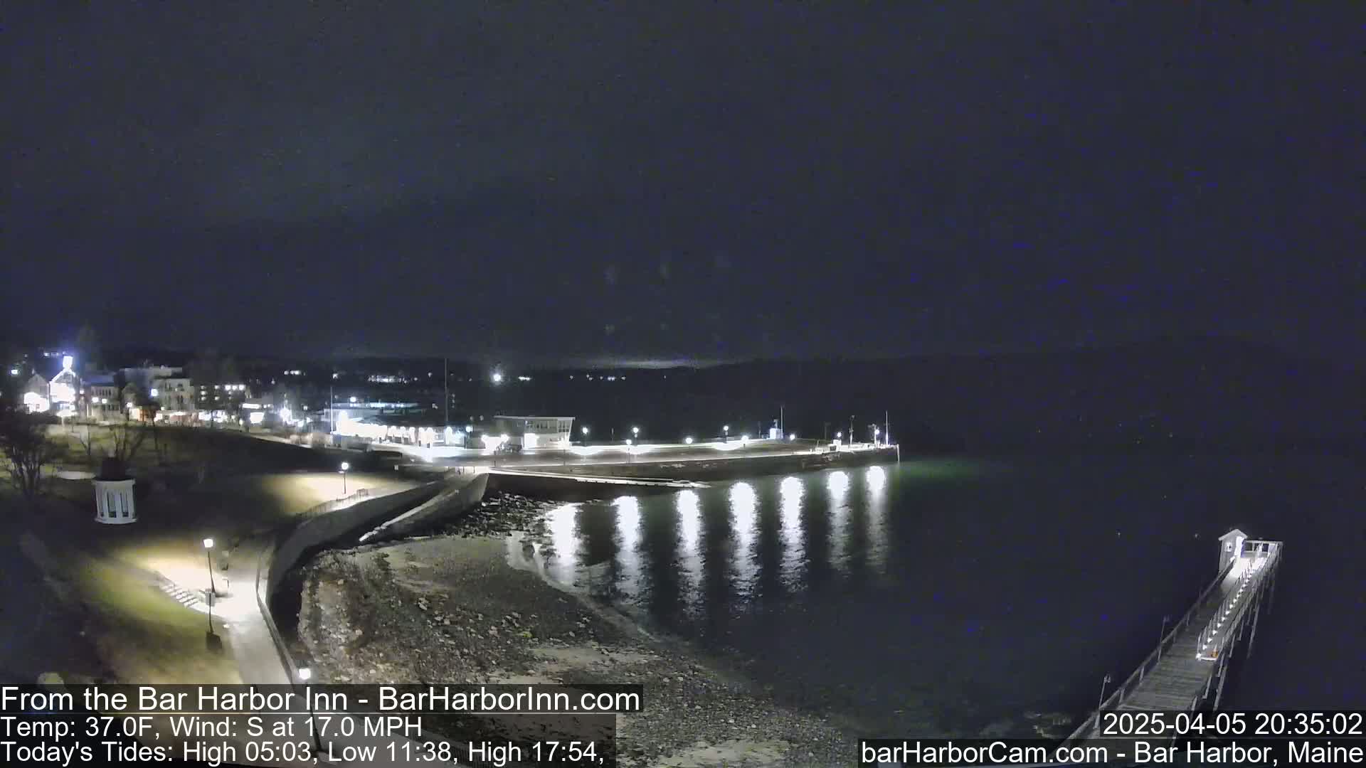A nighttime view of a calm, dark harbor with lights illuminating a shoreline and a pier extending into the water.