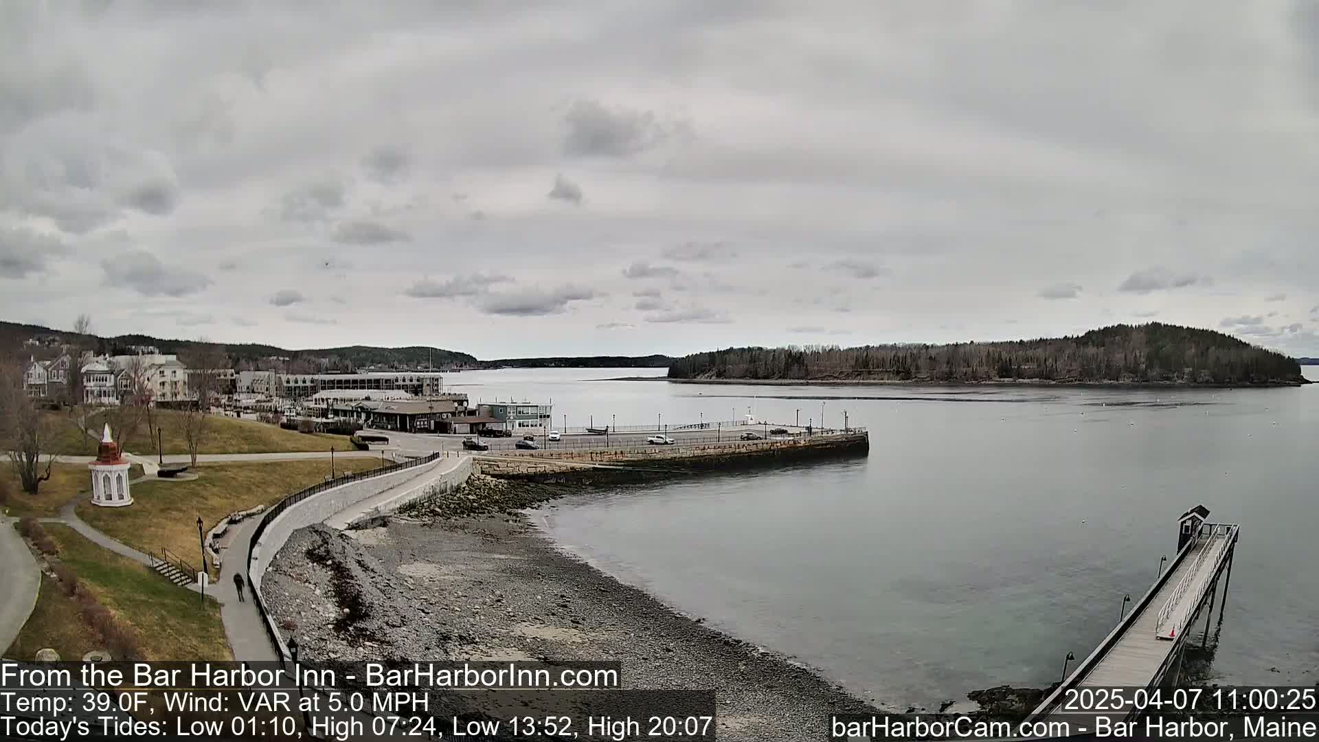 A mostly cloudy day reveals a coastal town with buildings, a park, and a long pier extending into calm water, with a small island in the distance.