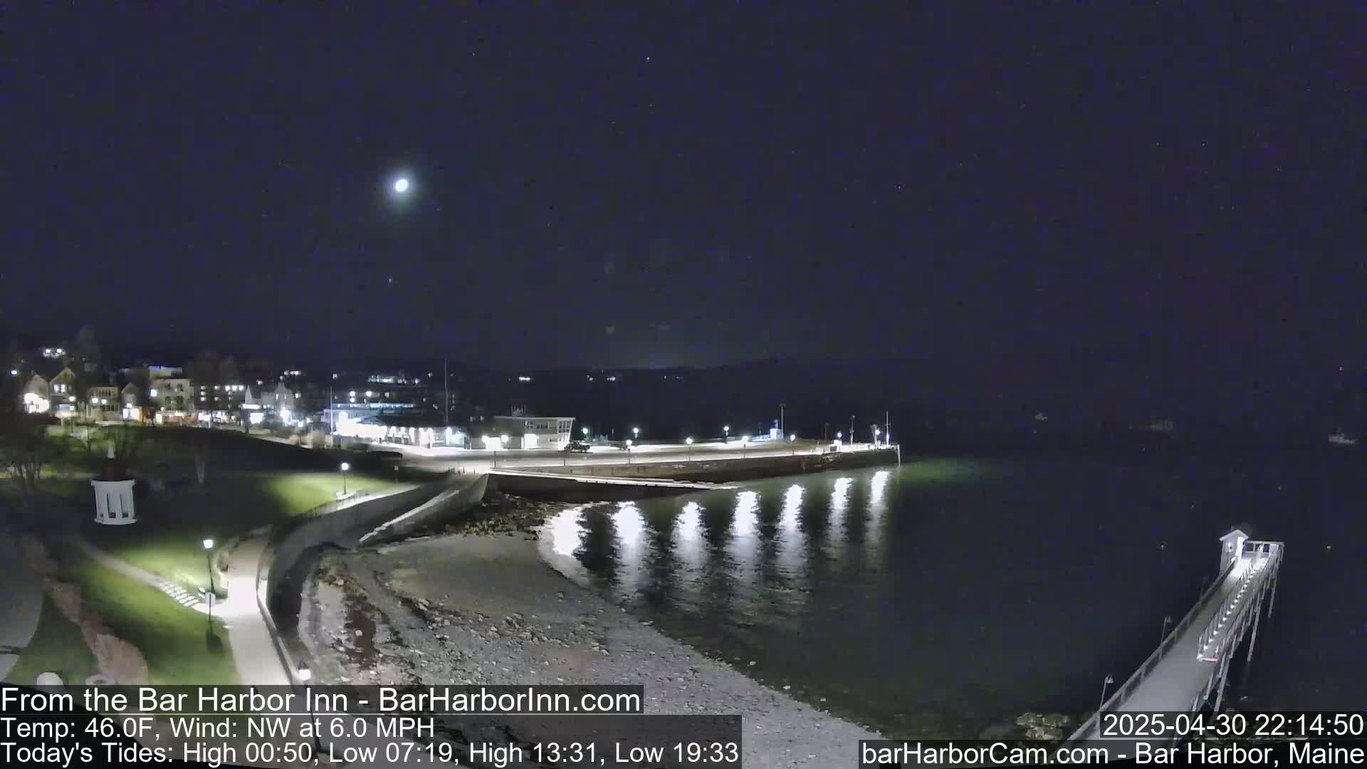 A nighttime coastal scene shows a town, a shoreline with a pier extending into calm water under a clear, moonlit sky.