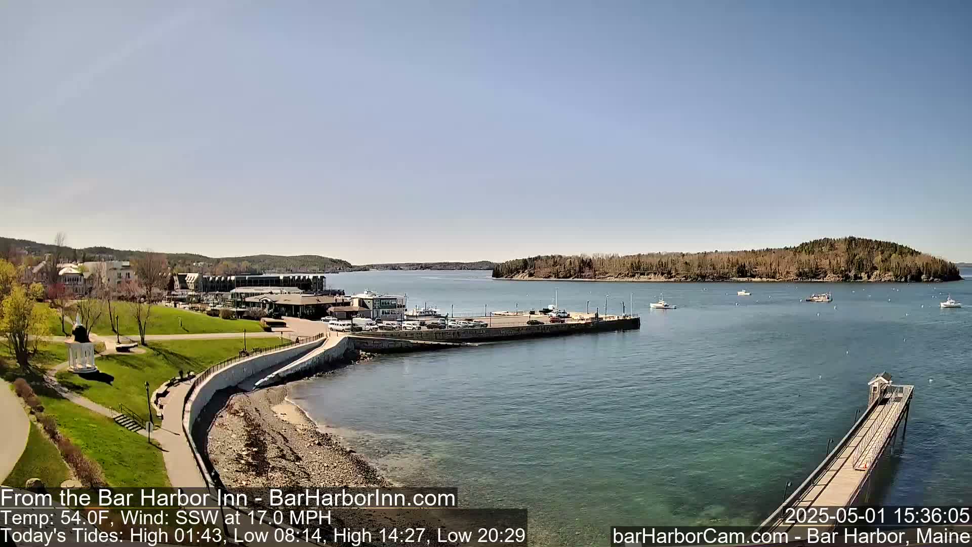A coastal town's harbor scene on a sunny day shows a calm body of water with several boats, a long pier, and green parkland with buildings in the background.