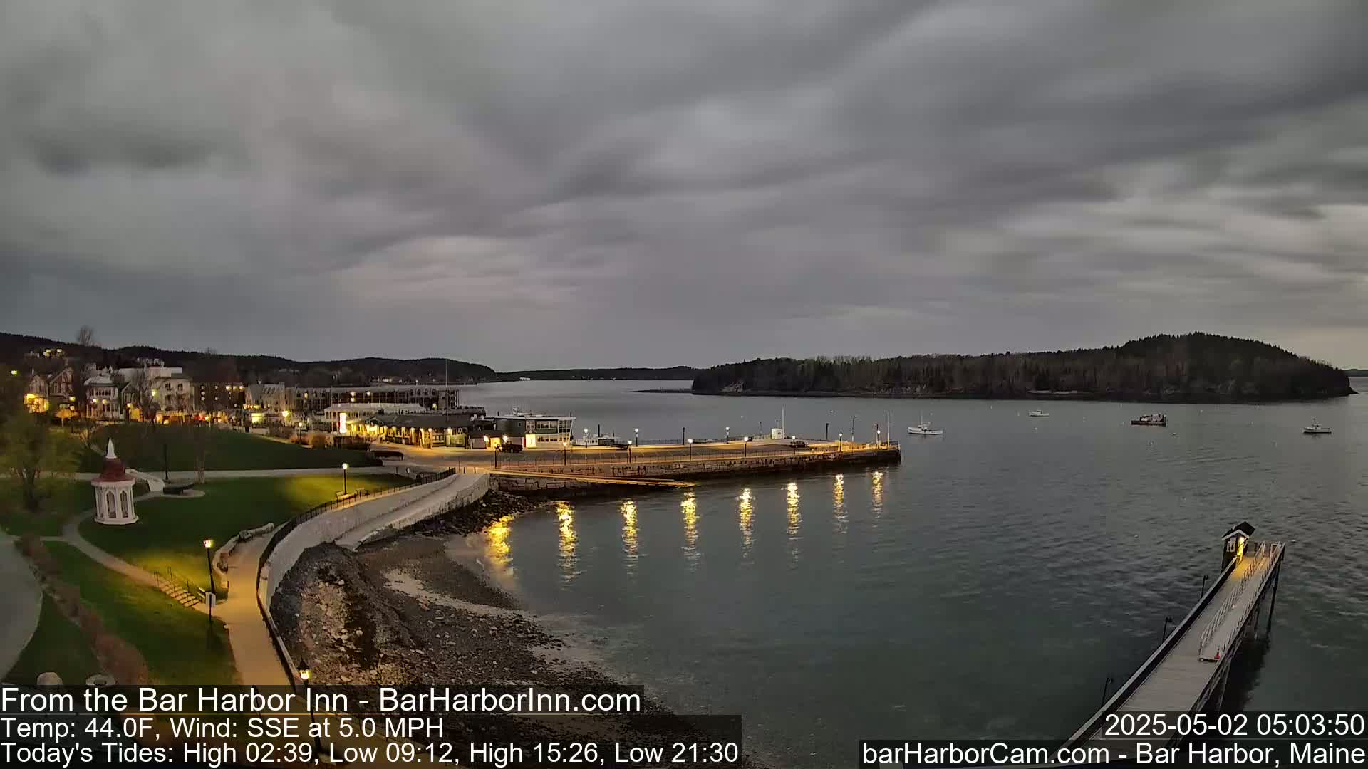 A coastal town at dusk under an overcast sky, with calm water reflecting lights from a waterfront area and pier.