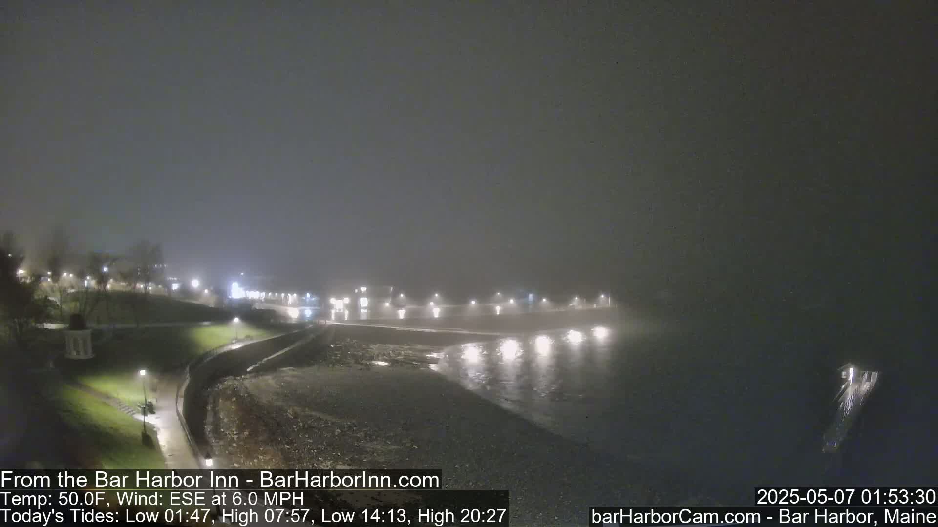 A foggy nighttime coastal scene shows a dark beach and calm water under a dim sky, with lights visible in the distance along a shoreline.