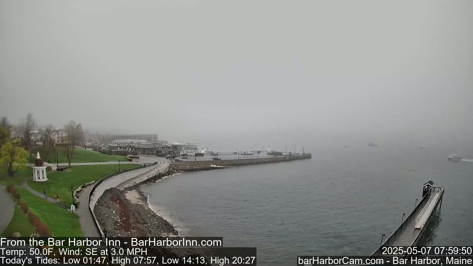 A foggy waterfront scene shows a calm body of water with several boats, a long pier extending into the water, and a grassy area with buildings in the background.