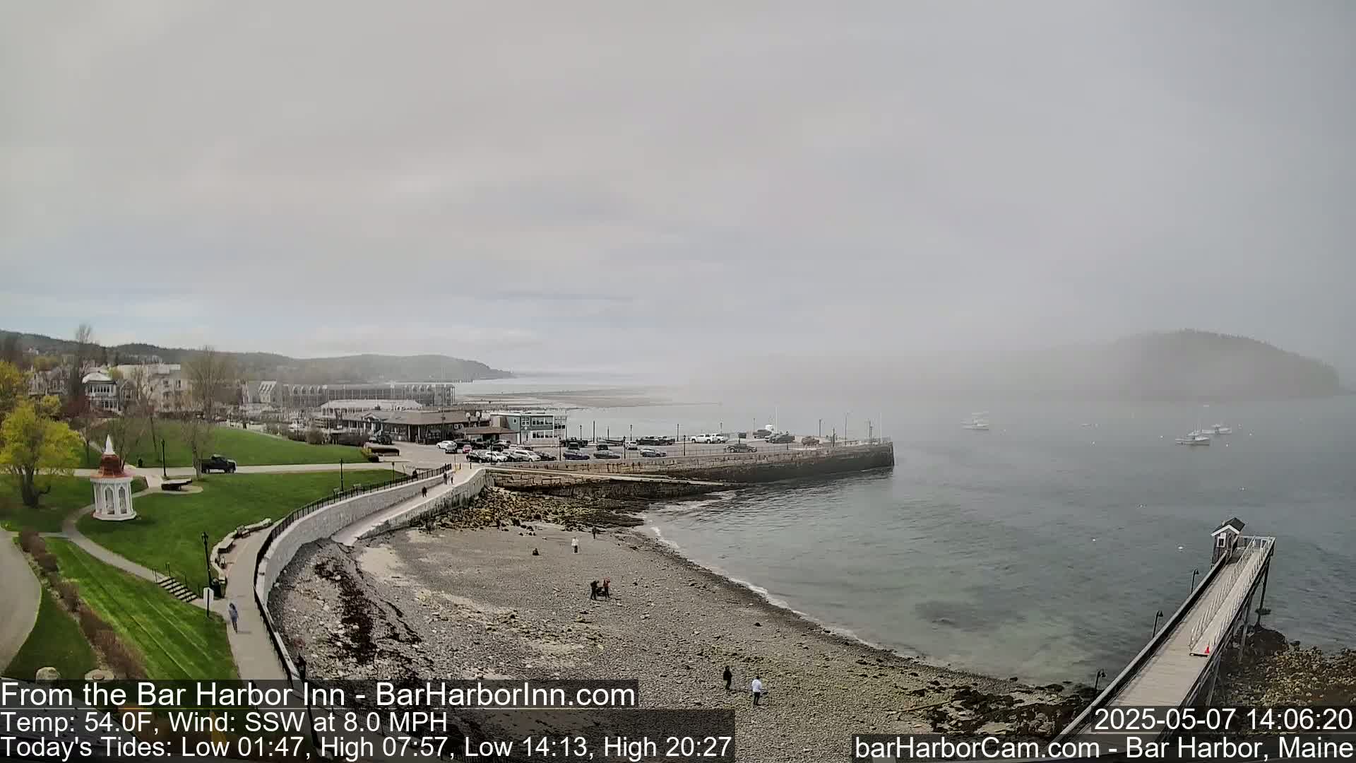 A foggy coastal scene shows a rocky beach, calm water, a long pier, and buildings in the background.