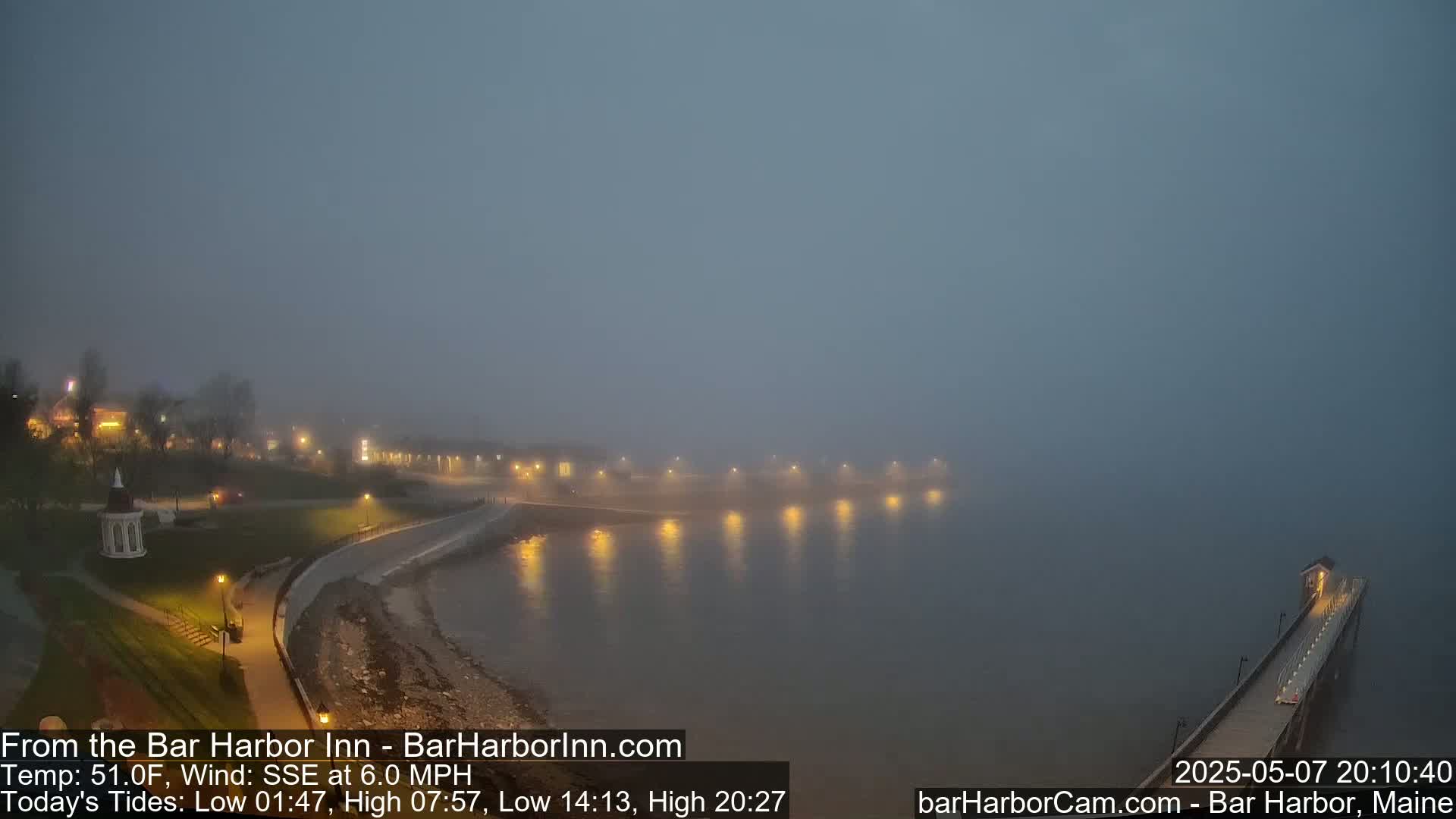 A foggy night scene shows a shoreline with lights illuminating a paved walkway, buildings, and a long pier extending into calm, misty water.