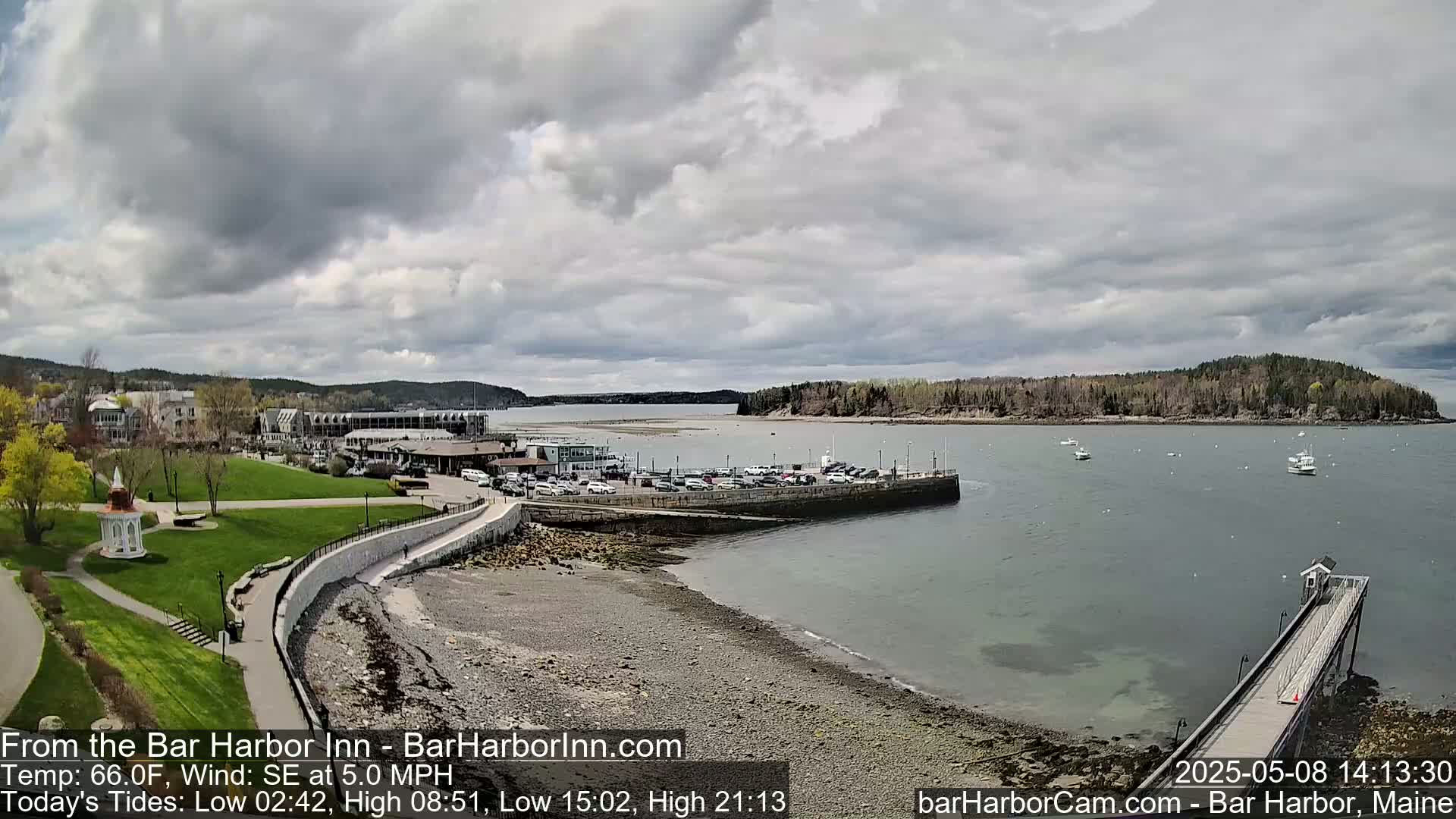 A harbor scene under a cloudy sky shows a town, a grassy park, a parking lot, a rocky shoreline, a pier, and several boats on the water.