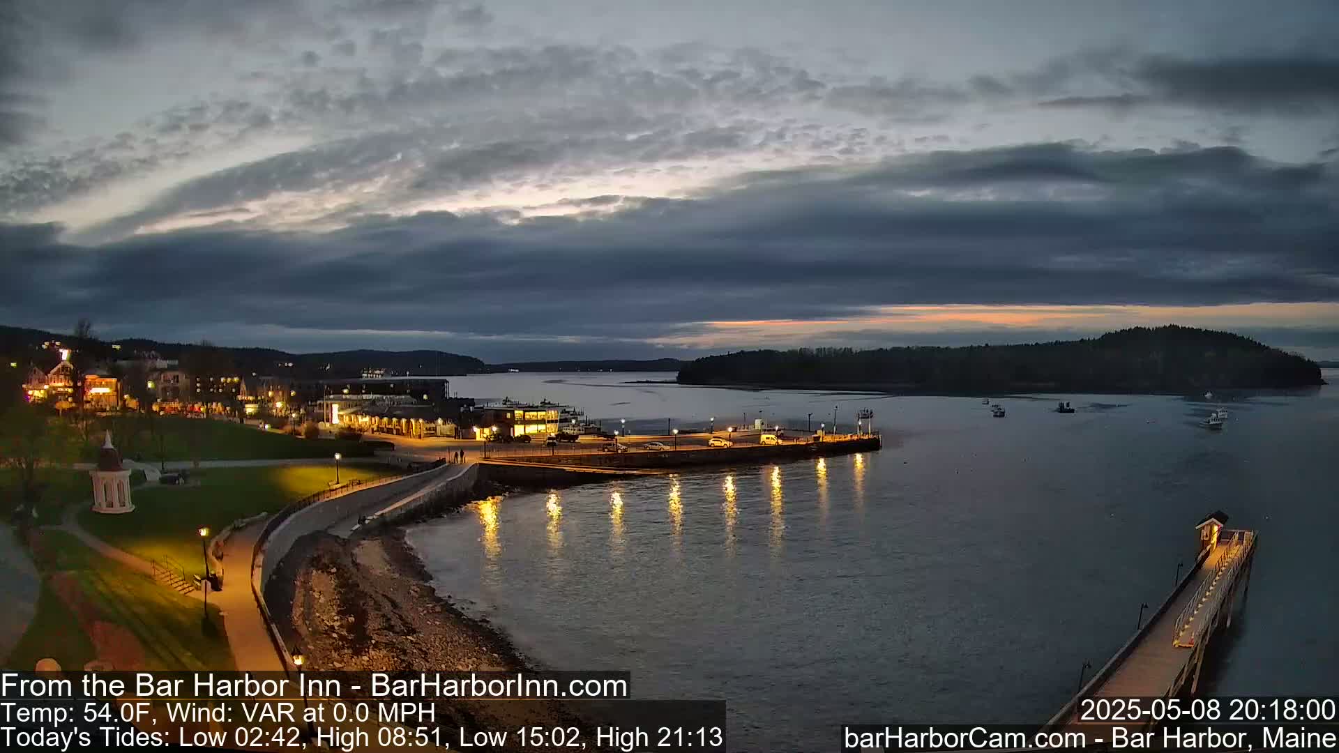 A harbor town at twilight under a mostly cloudy sky, with a calm body of water reflecting lights from a pier and buildings along the shore.