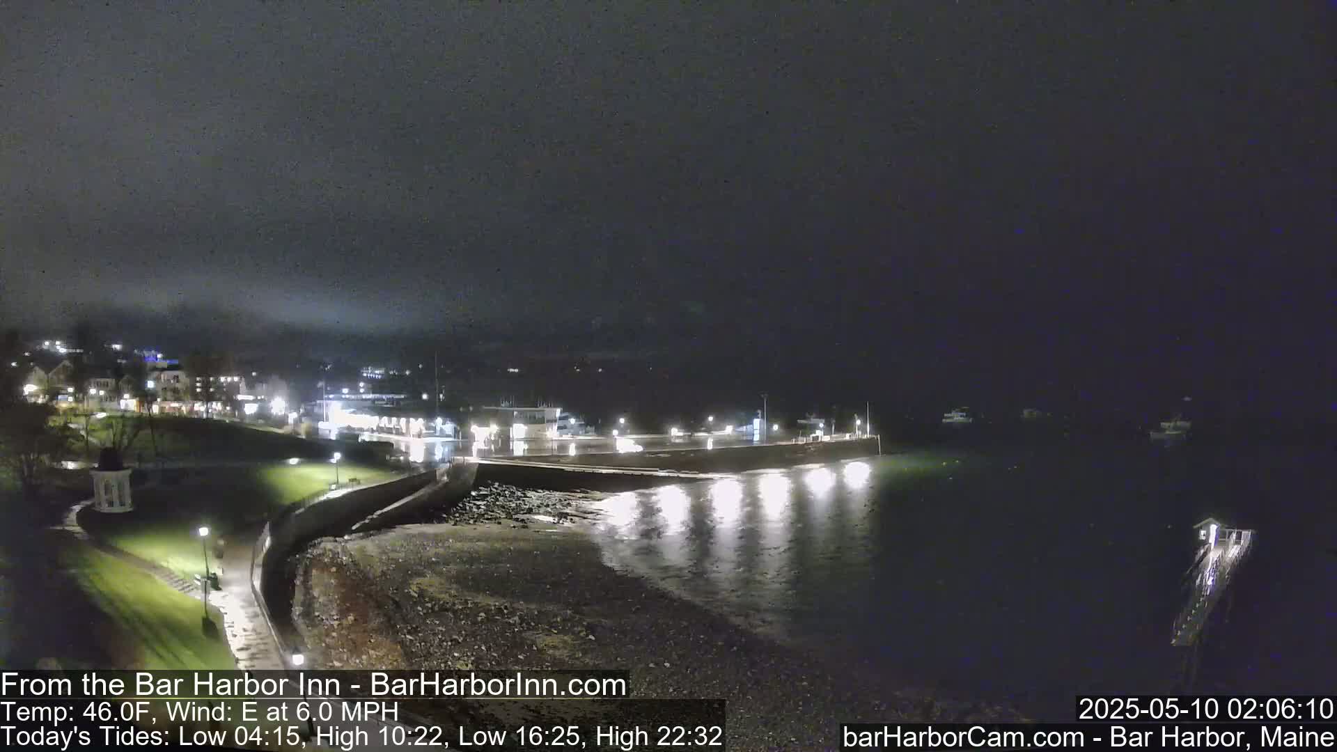 A nighttime coastal scene shows a dark, calm body of water reflecting lights from a nearby town under a foggy sky.