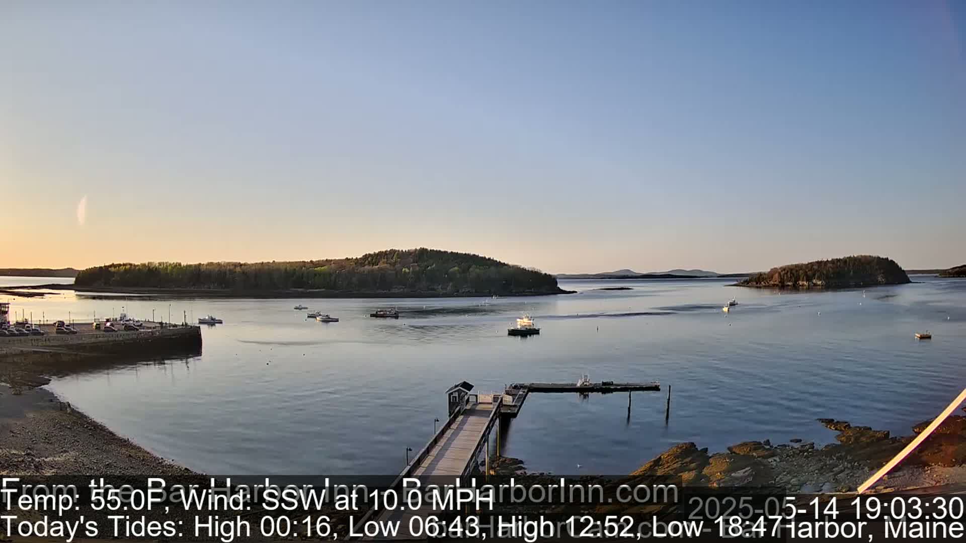 A calm harbor with several small boats, a long dock extending from a rocky shore, and forested islands in the background under a clear, twilight sky.