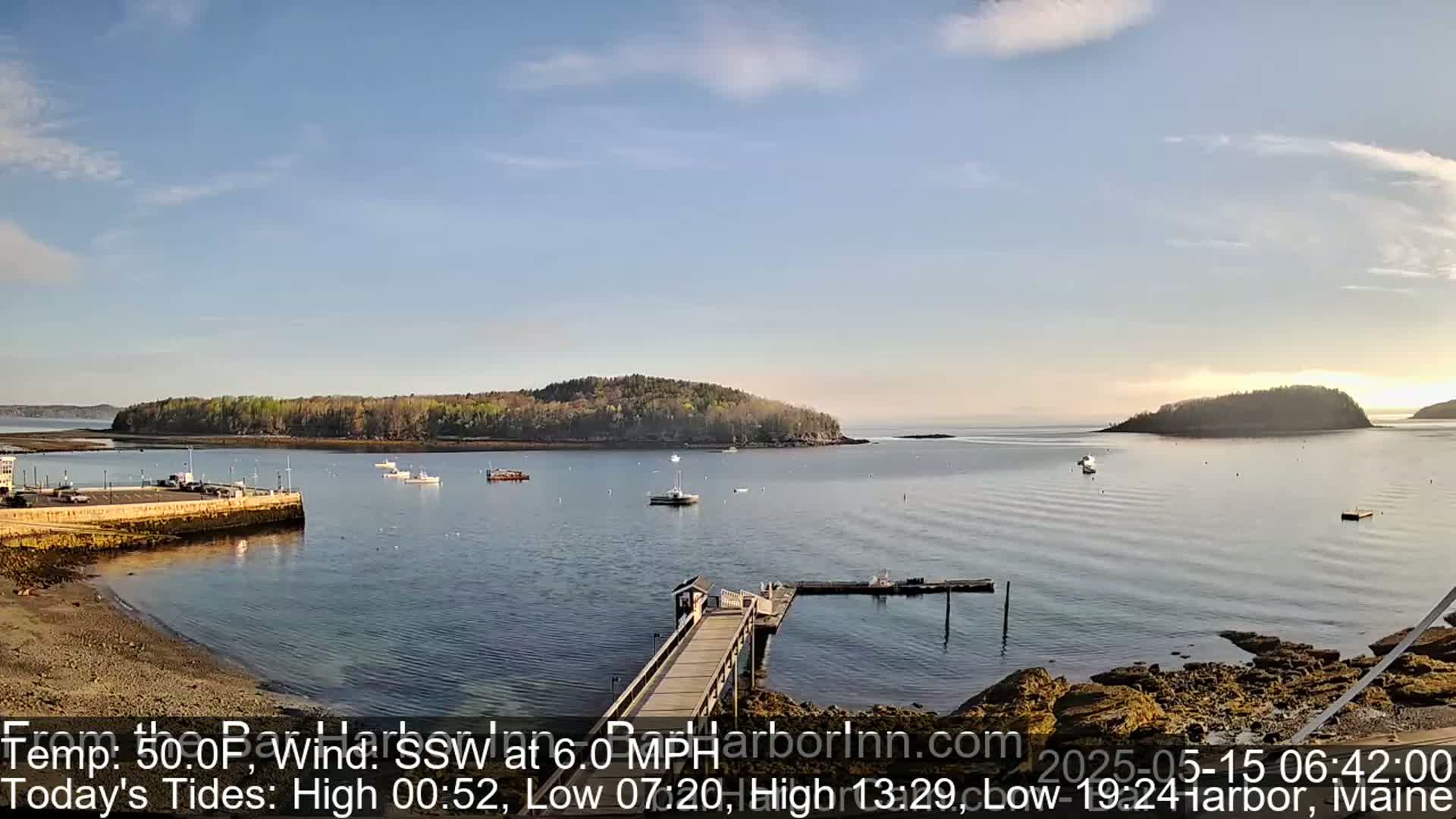 A calm, sunny day reveals a tranquil harbor scene with a long pier extending into the water, several boats scattered across the bay, and a small, tree-covered island in the distance.