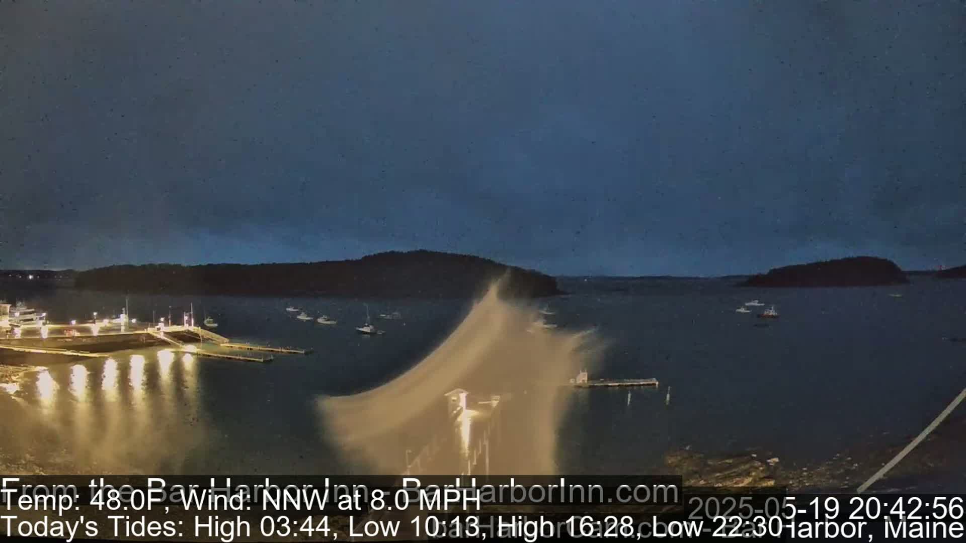A nighttime harbor scene shows a body of water with several boats and a pier, under a mostly clear, dark sky with a strong wind blowing sand or spray from the water near the pier.