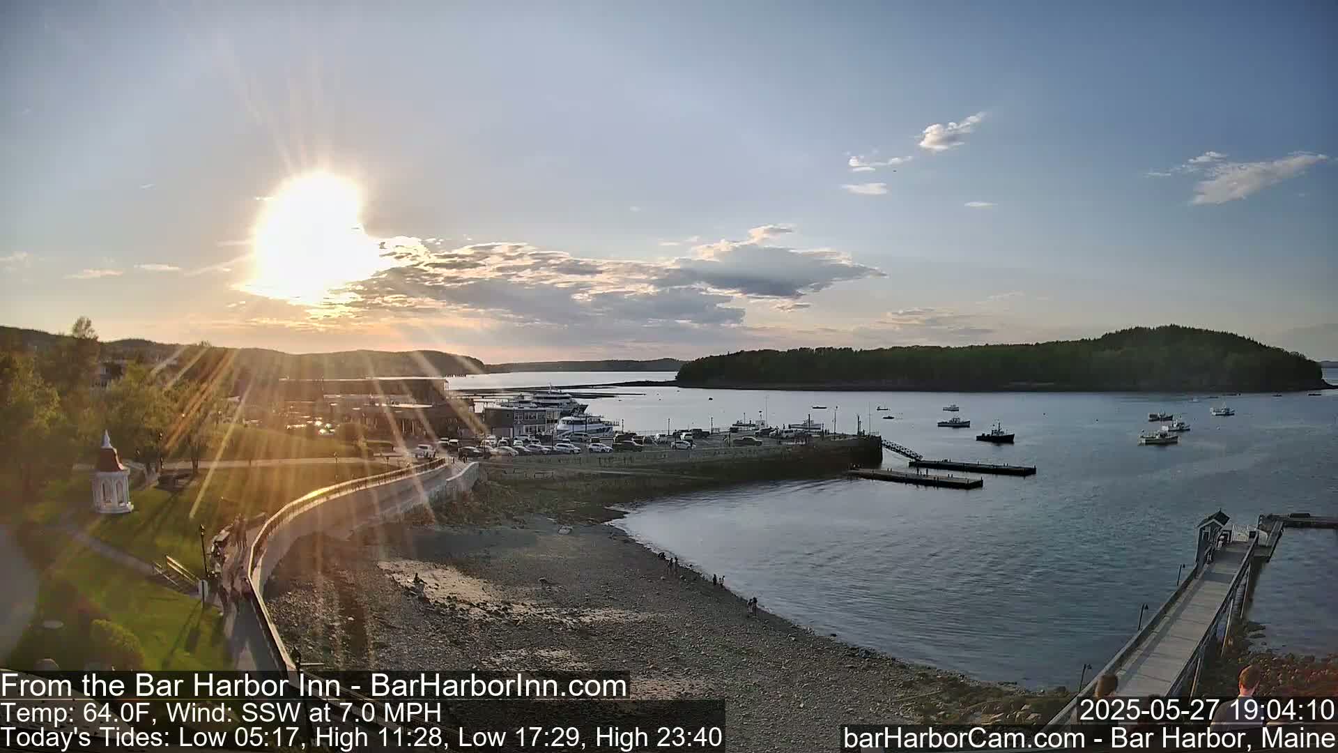 A sunny sunset over a calm harbor with boats moored at a pier, a town in the background, and a rocky shoreline in the foreground.