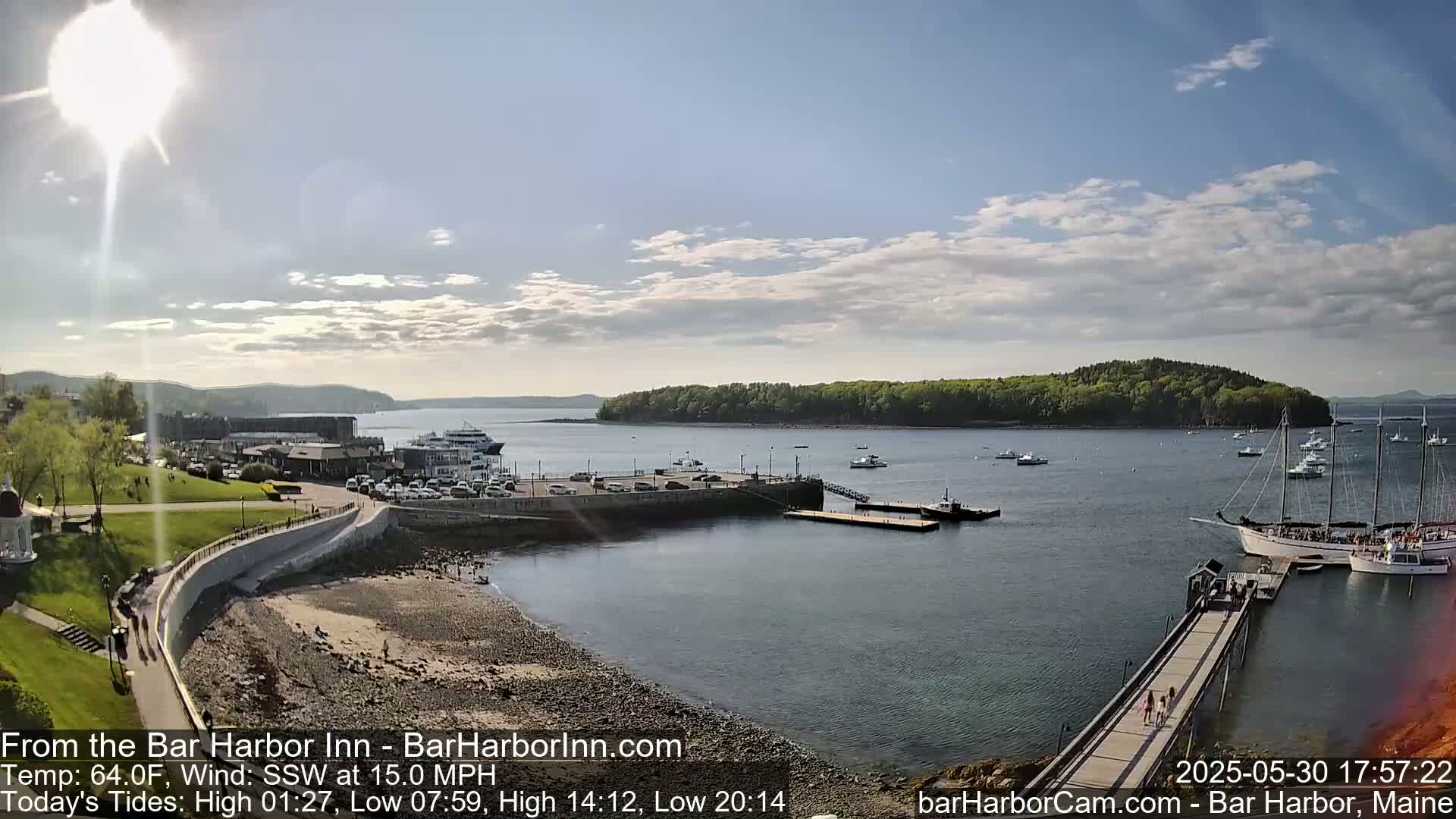 A sunny harbor scene with a partly cloudy sky shows a pier with several boats, including a large sailing vessel, and a ferry docked near a shoreline with people walking along a path.
