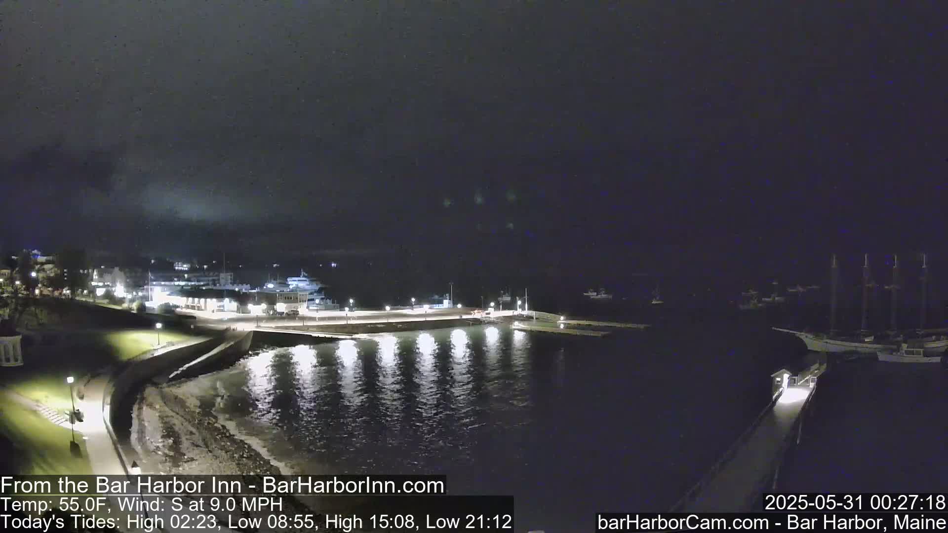 A nighttime view of a harbor with boats docked, lights illuminating the water and shoreline under a dark sky.