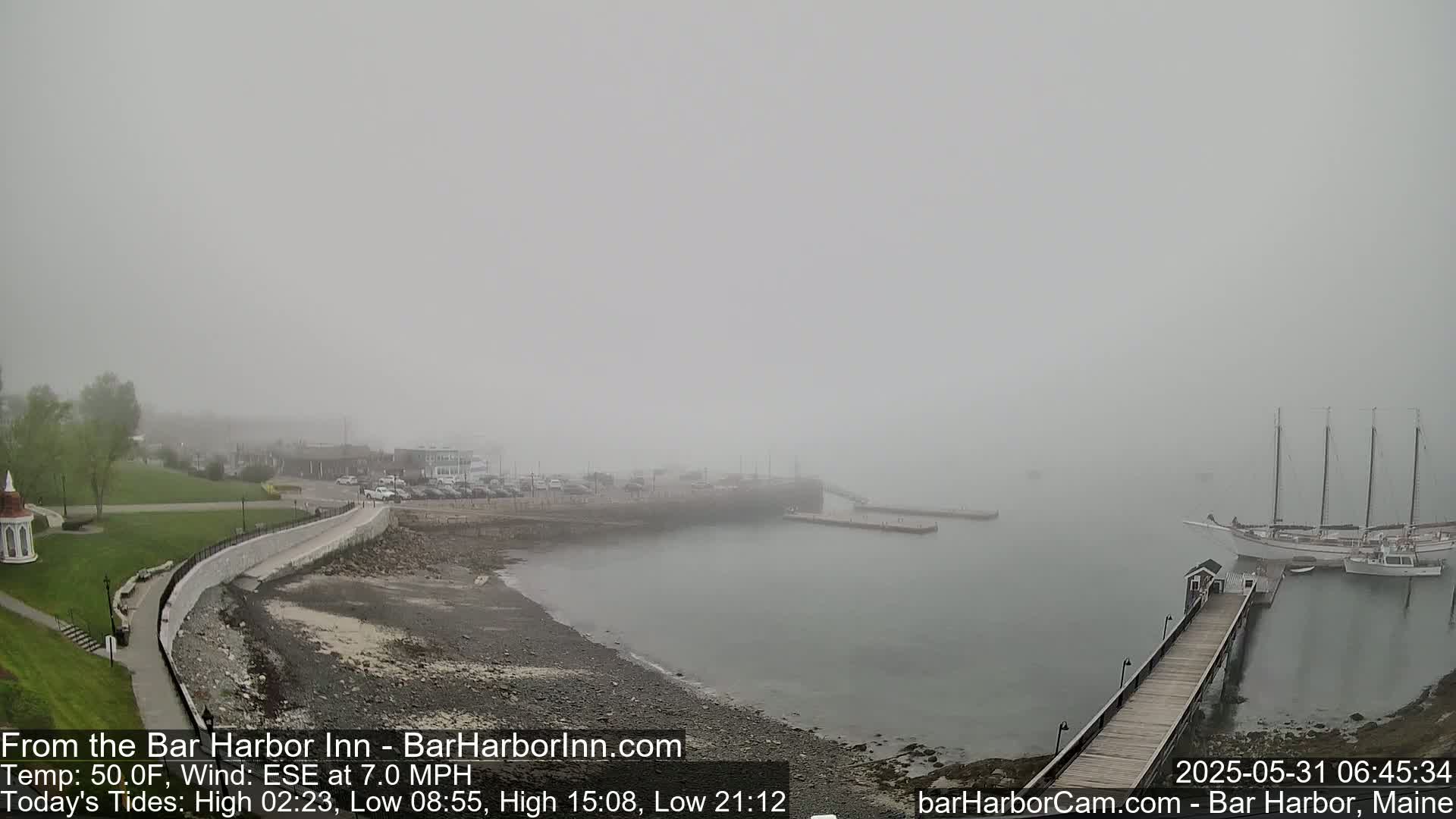 A foggy harbor scene shows a rocky shoreline, a wooden walkway leading to a pier with a large sailboat moored nearby, and a calm body of water extending to a distant, indistinct coastline.