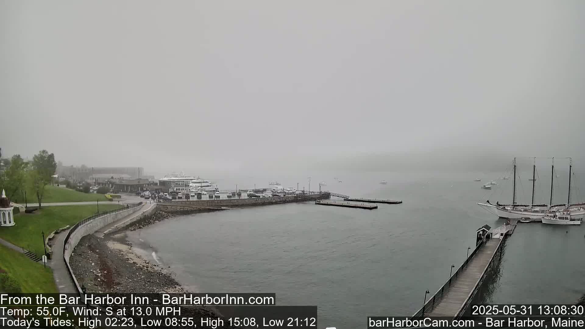 A foggy harbor scene shows a town, boats docked at a pier, and a large sailboat in the water.