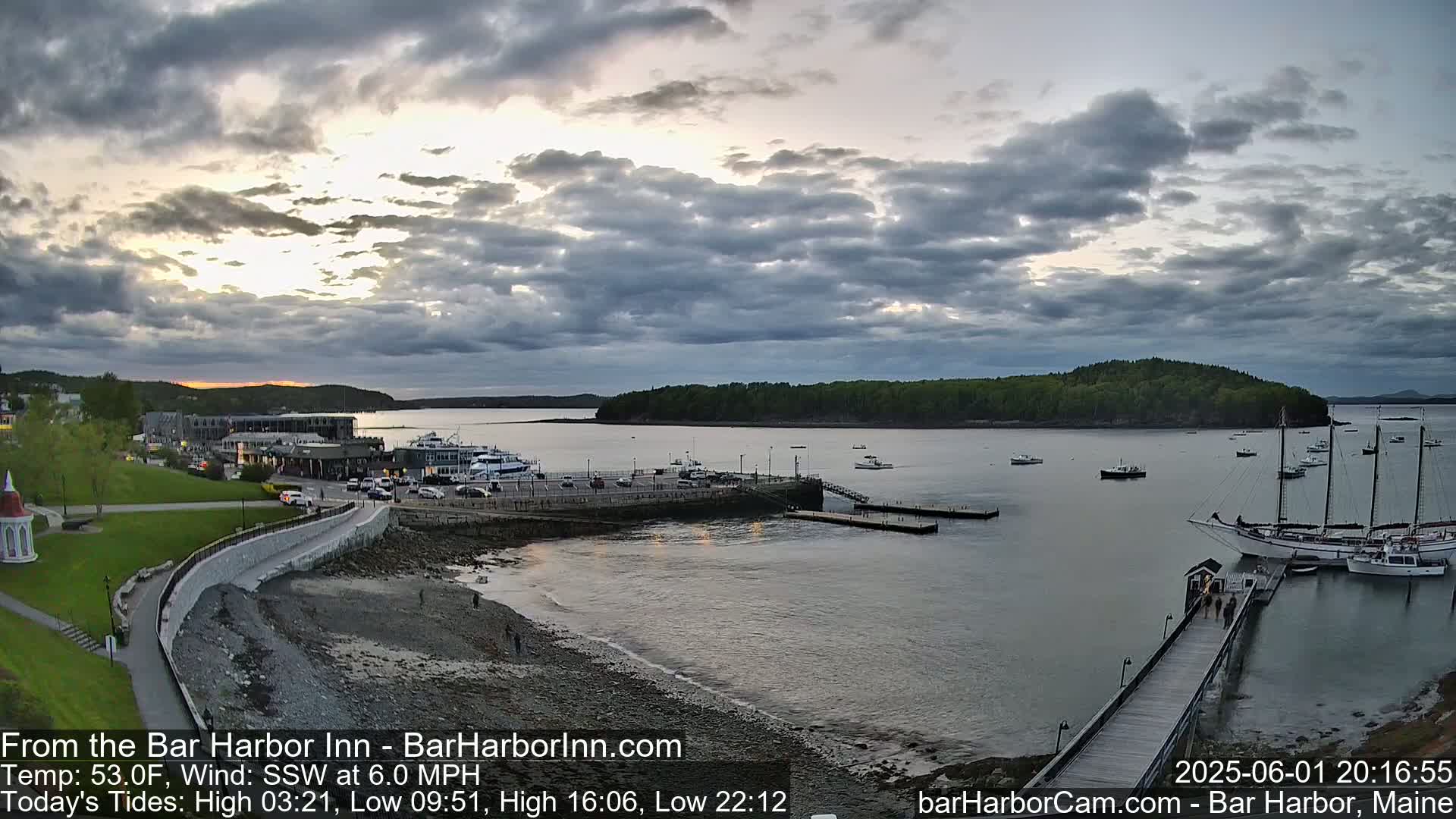 A harbor scene at sunset under a partly cloudy sky shows boats moored at a pier with a grassy area and buildings in the background.