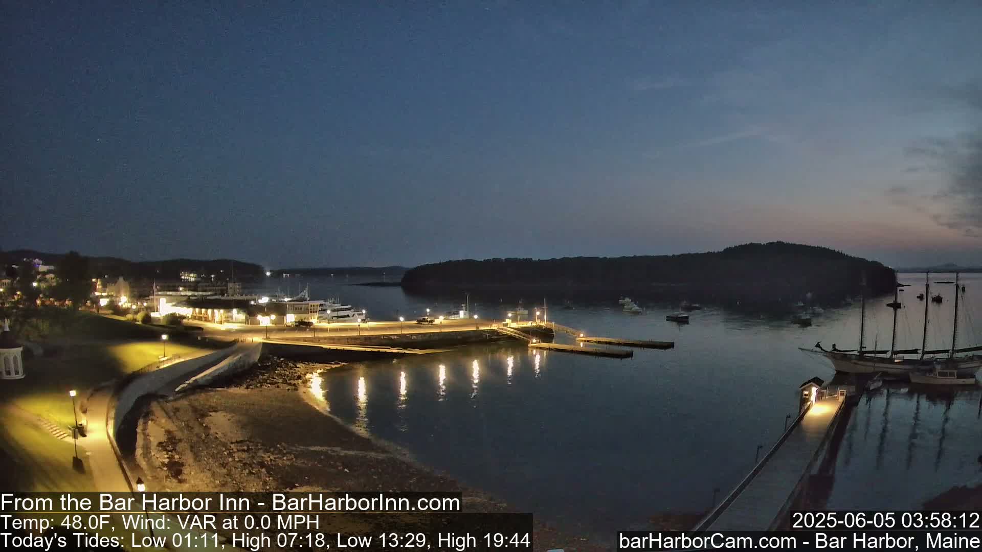 A nighttime view of a harbor with boats moored at a dock, illuminated buildings along the shoreline, and calm water under a twilight sky.