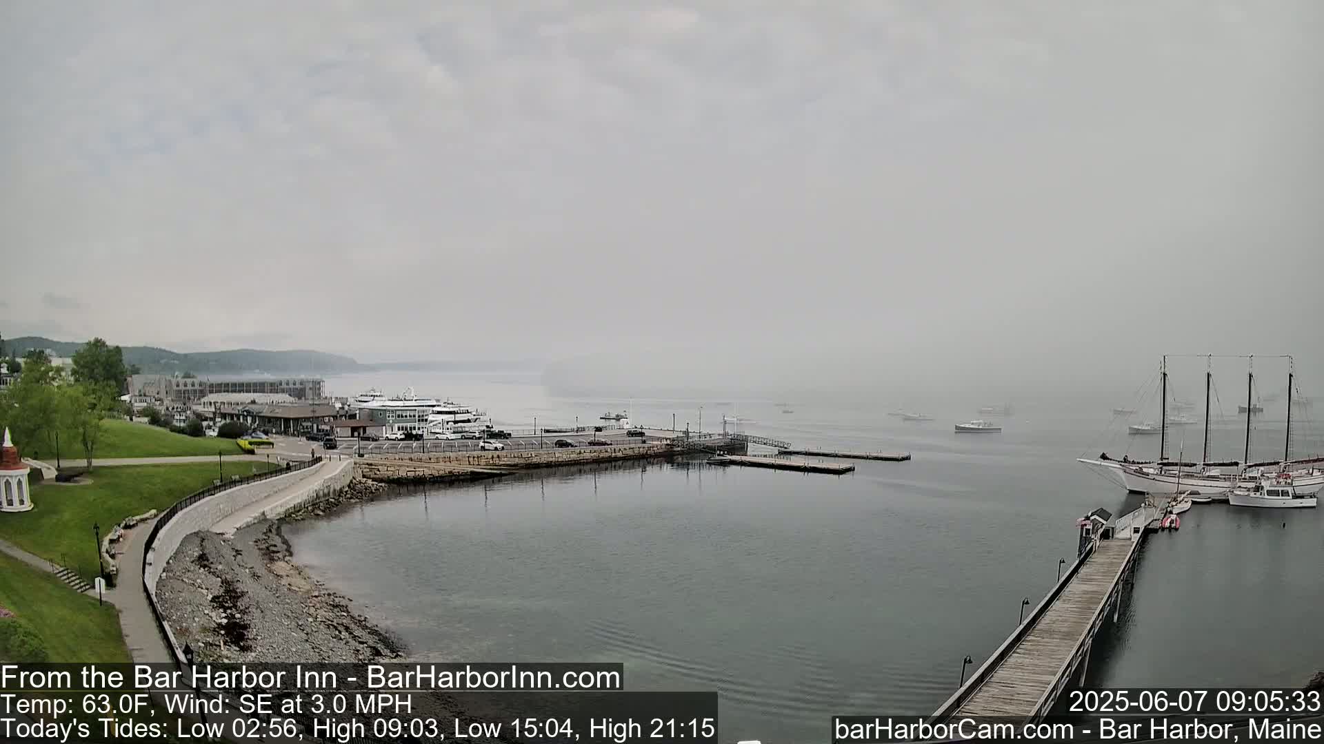 A foggy harbor scene shows a waterfront with docked boats and ships, a long pier extending into the water, and a grassy area with buildings in the background.