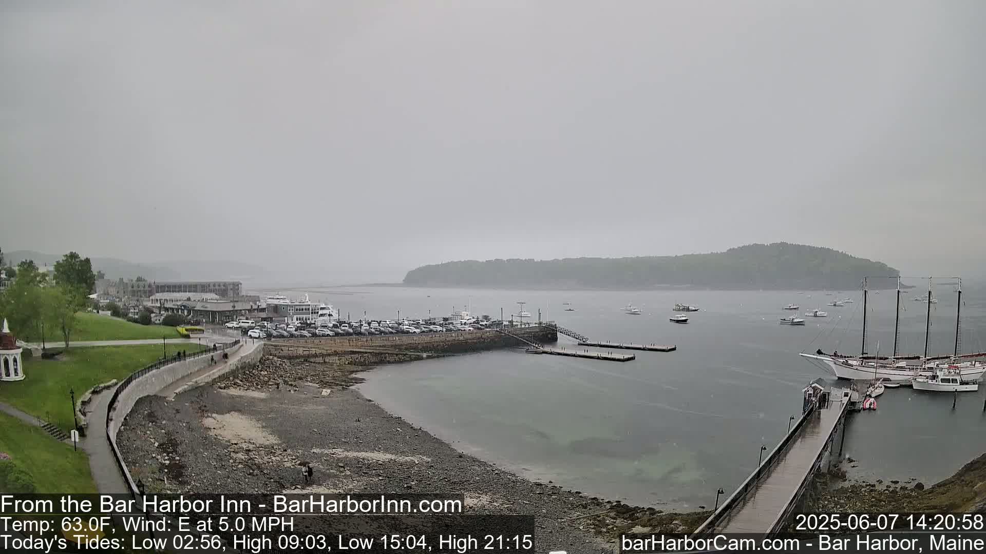A harbor scene on an overcast day shows a rocky shore with a parking lot and buildings in the background, several boats in the water, and a long dock extending into the harbor.