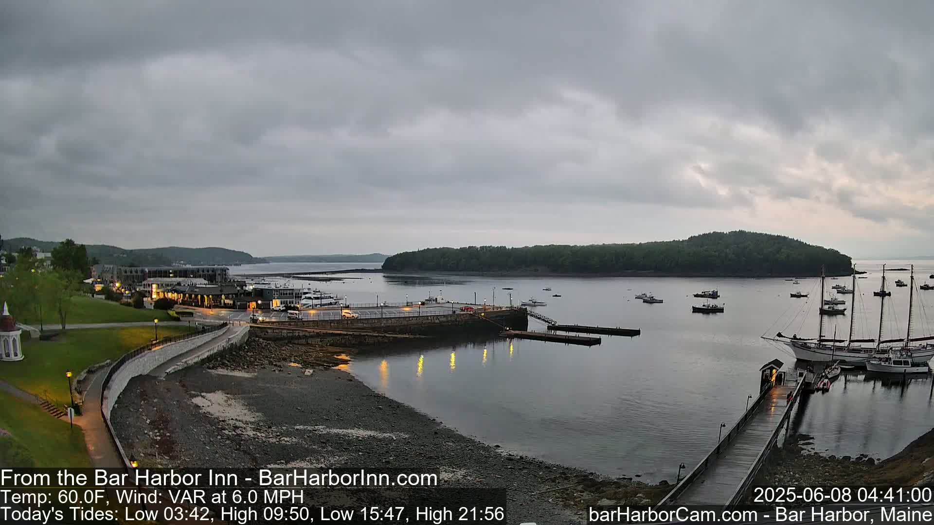 A calm harbor scene under an overcast sky shows a shoreline with buildings, a paved walkway, a rocky beach, and several boats moored at a dock.
