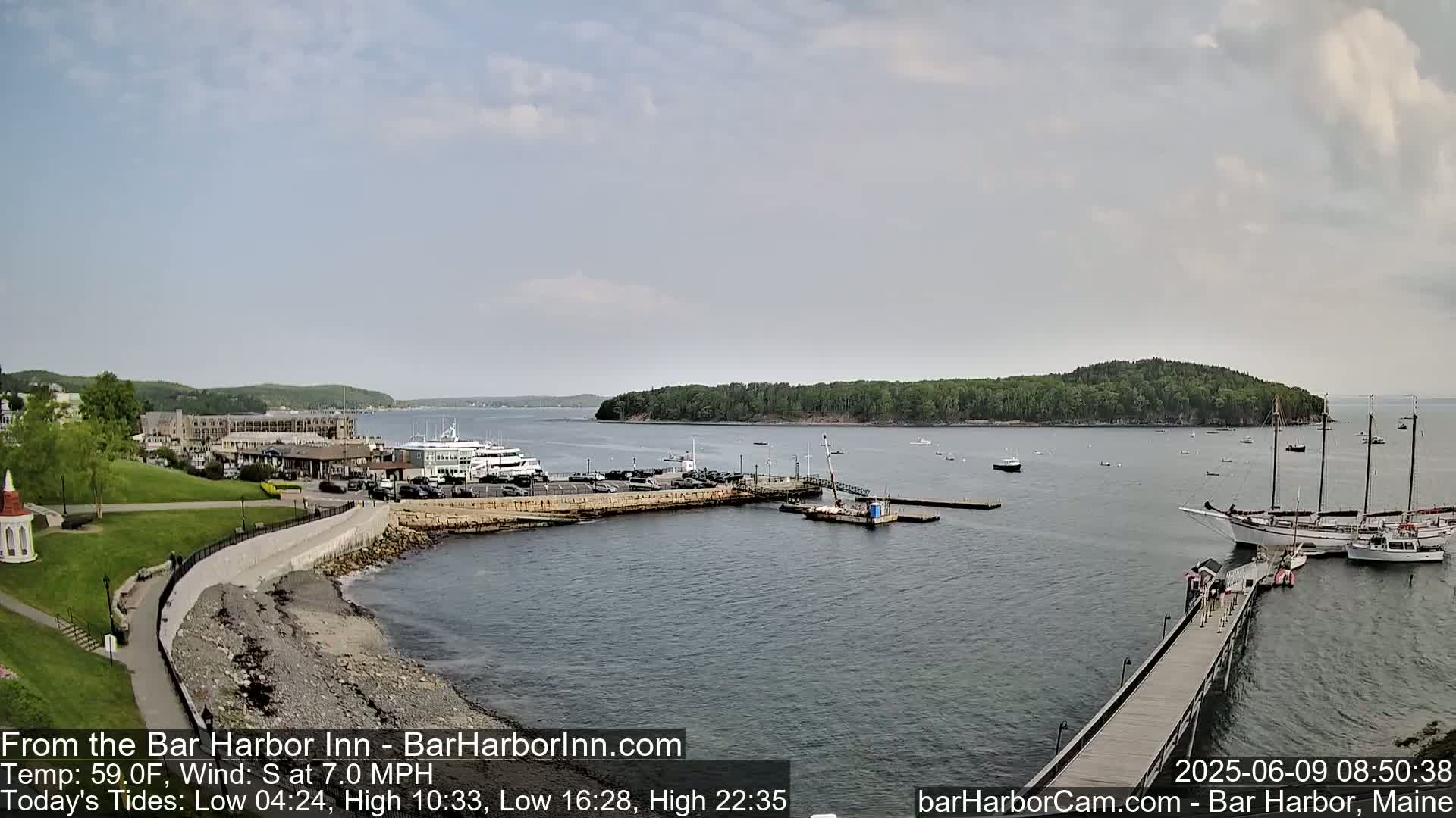 A harbor scene on a partly cloudy day shows a parking lot, buildings, boats, and a long pier extending into the water.
