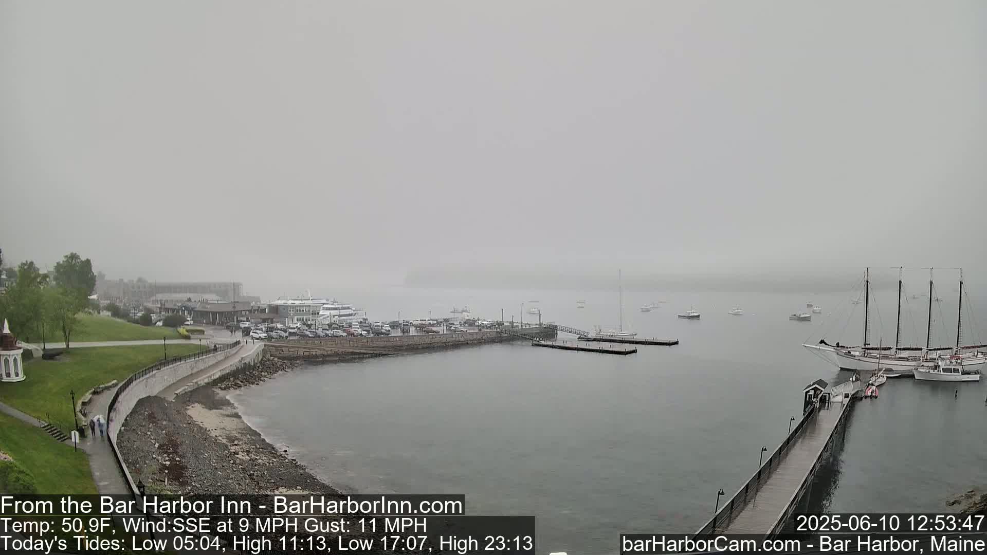 A foggy harbor scene shows a shoreline with a paved walkway, green grass, and buildings, with several boats and a large sailing ship moored in the water.