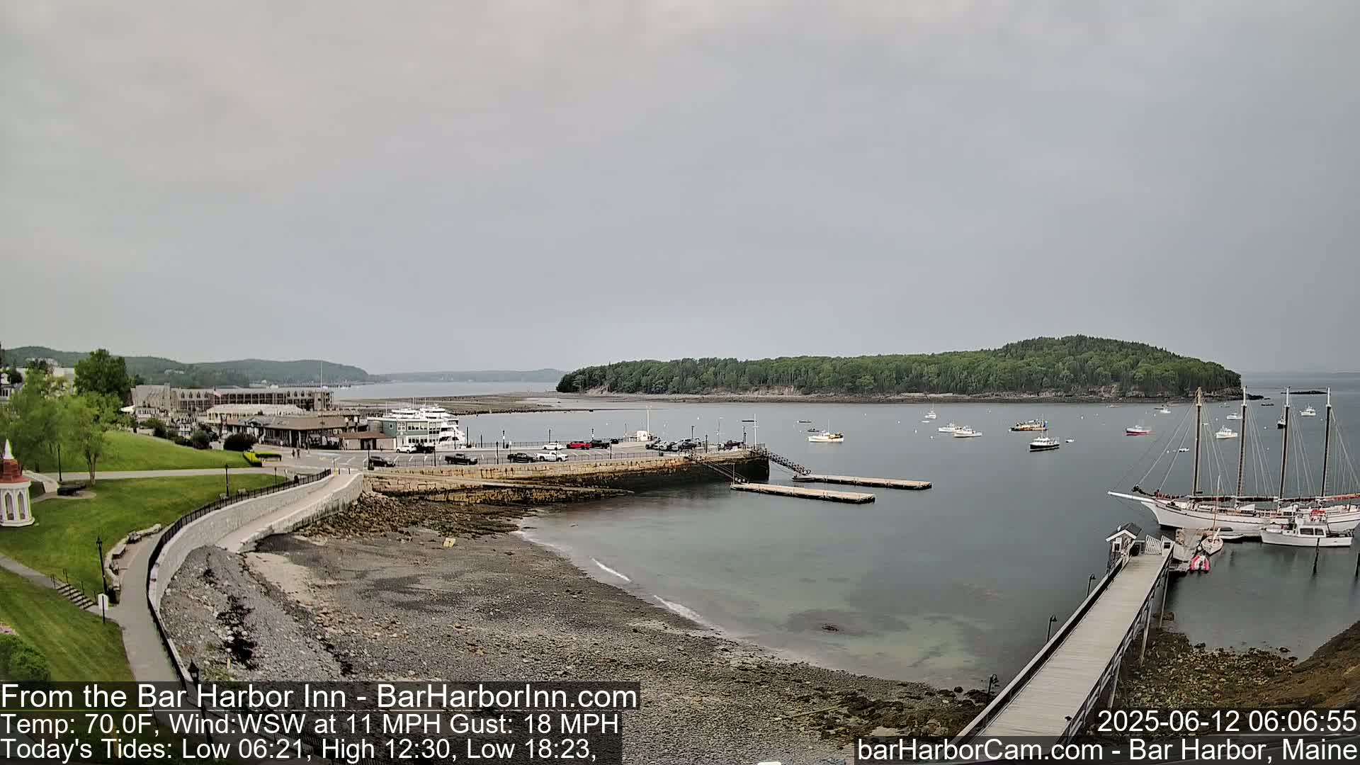 A calm harbor scene on an overcast day shows a shoreline with a parking lot, buildings, a walkway, a small dock, several boats, and a large sailboat at anchor.