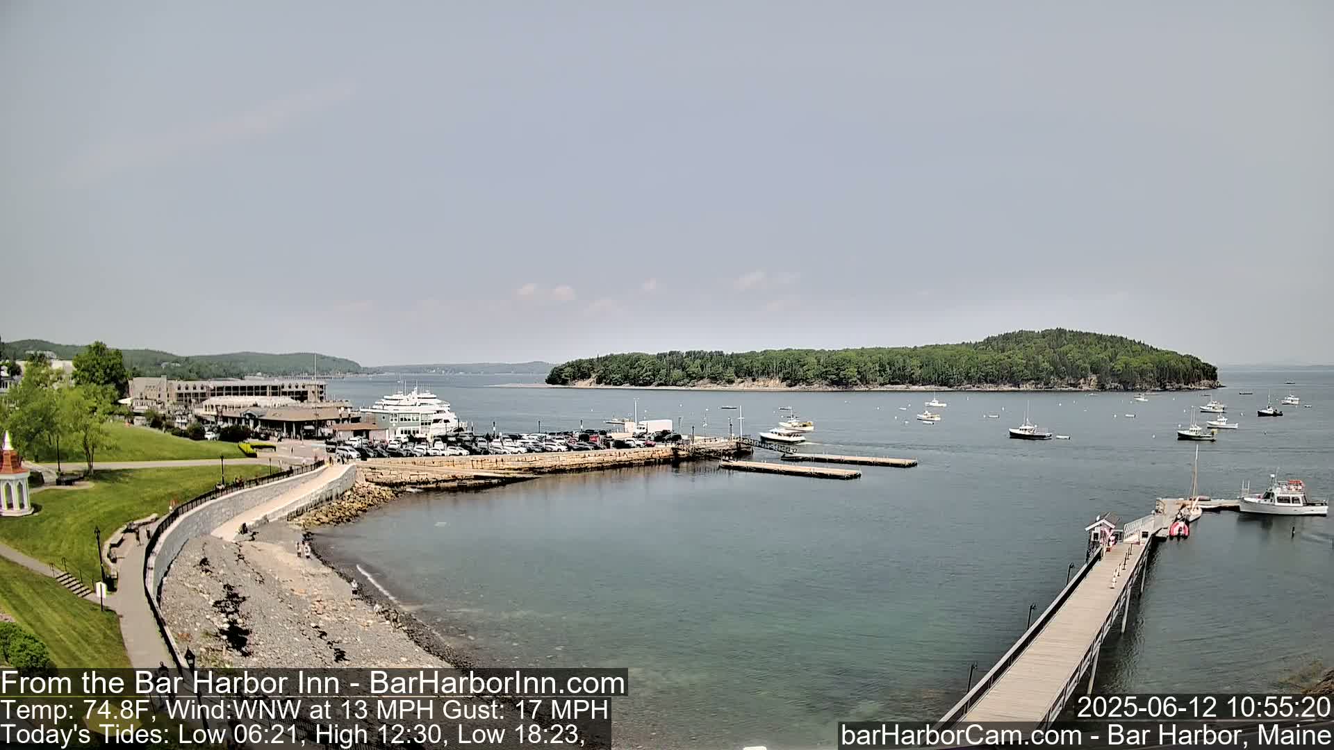 A harbor scene on a partly sunny day shows a calm body of water with numerous boats docked and at anchor near a stone seawall and a long pier extending into the water, with a large building and green area visible on the shore.