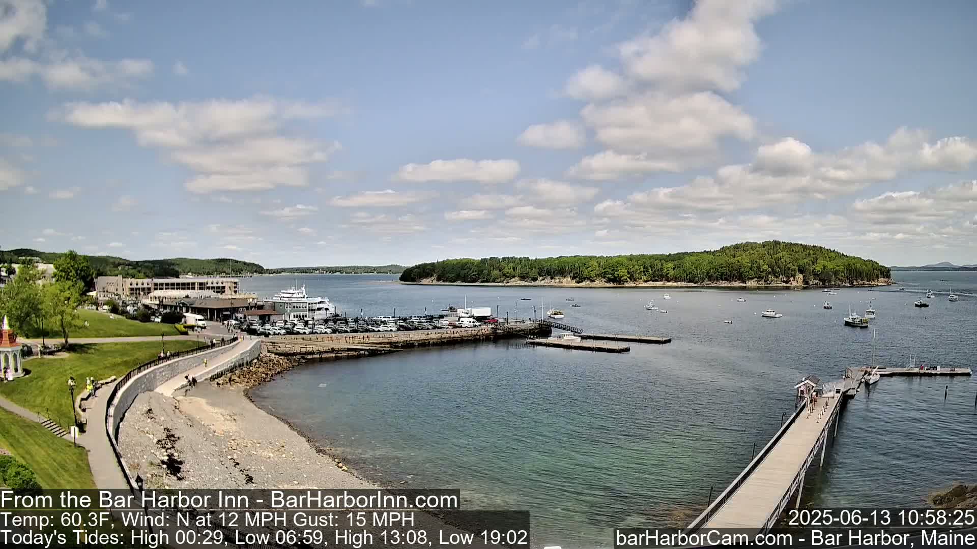 A harbor scene on a partly cloudy day shows a waterfront with boats, a parking lot, buildings, and a long pier extending into the calm water.