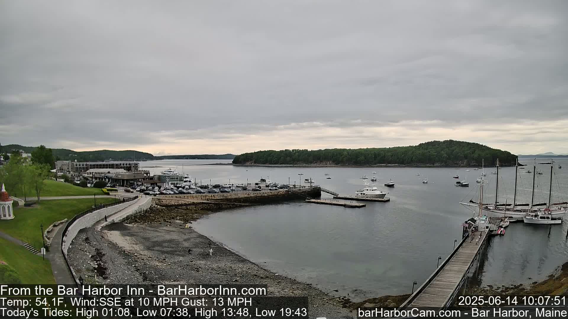 A harbor scene under an overcast sky shows a calm body of water with several boats moored at a pier, a parking lot, and green land beyond.