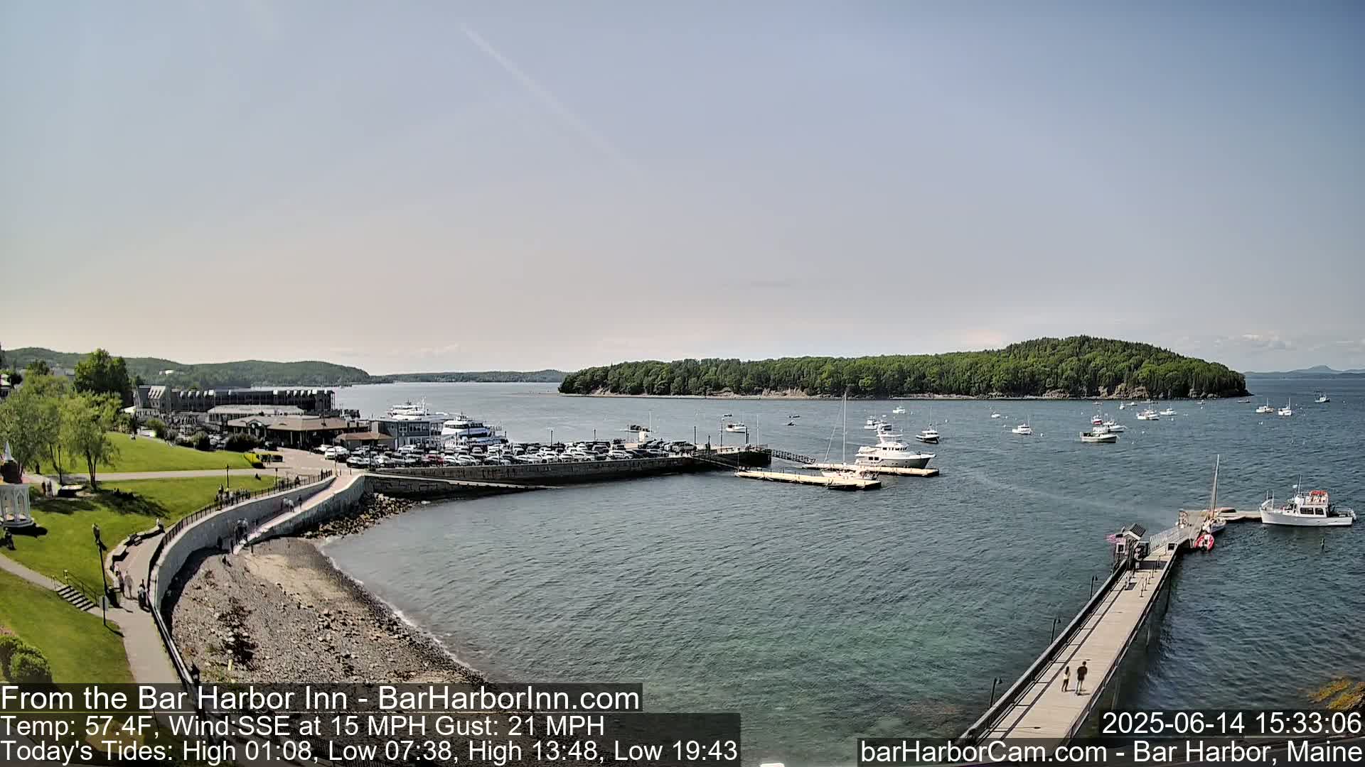 A harbor scene under an overcast sky shows a calm body of water with several boats moored at a pier, a parking lot, and green land beyond.