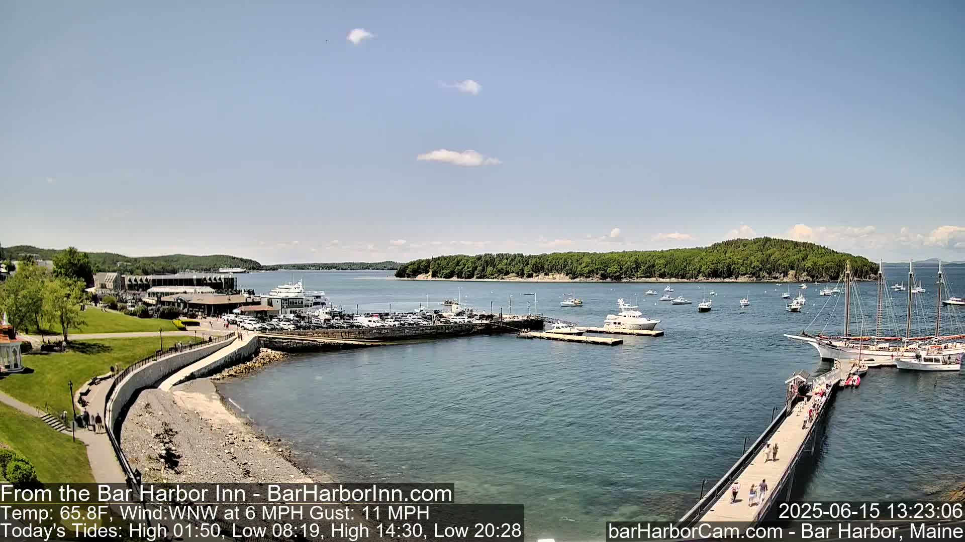 A harbor with numerous boats, a grassy area with buildings, and a long pier extending into the water on a partly sunny day with a light wind.