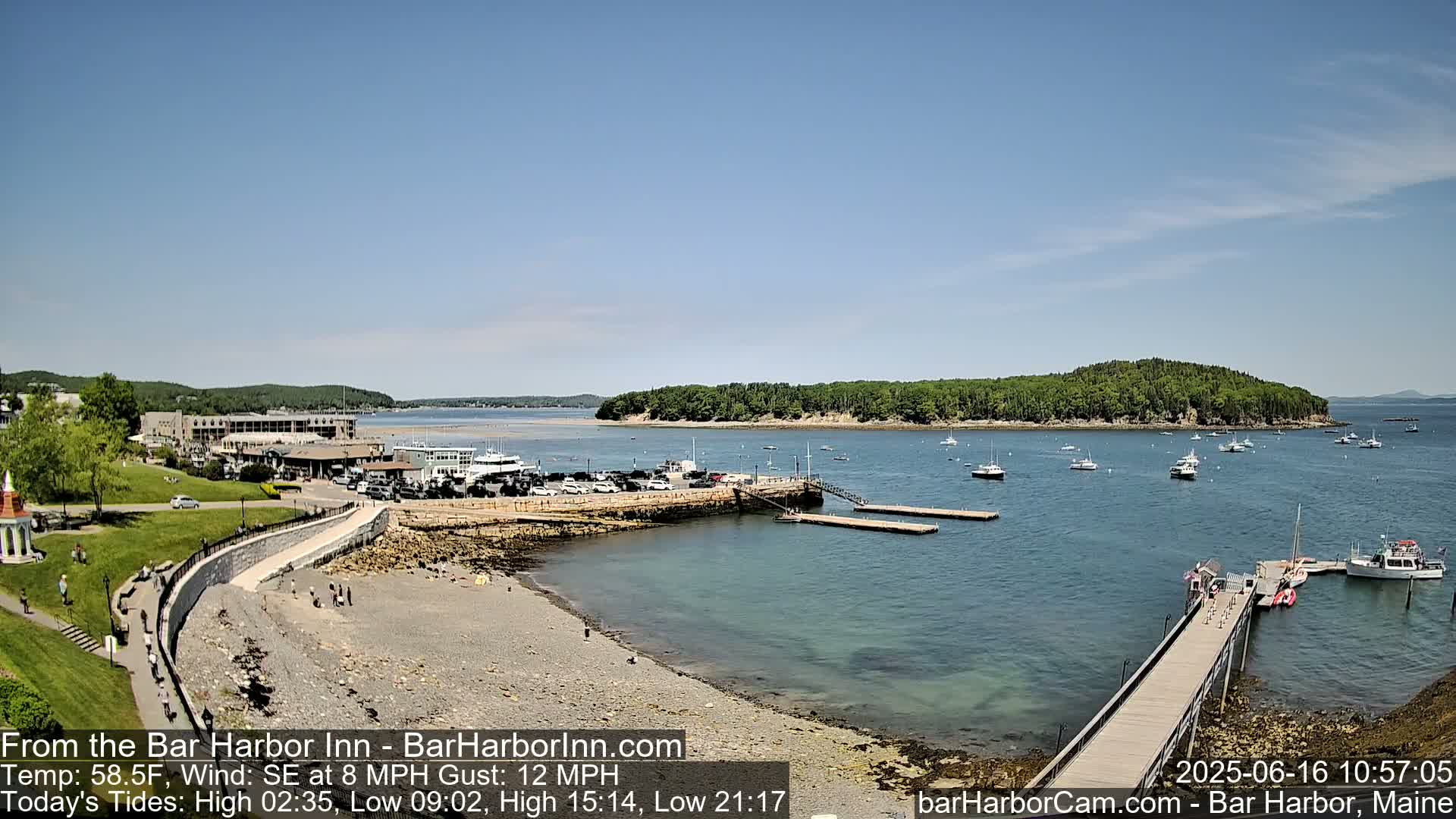 A sunny harbor scene shows a rocky shoreline with a pier, several boats, and a calm body of water with a wooded island in the distance.