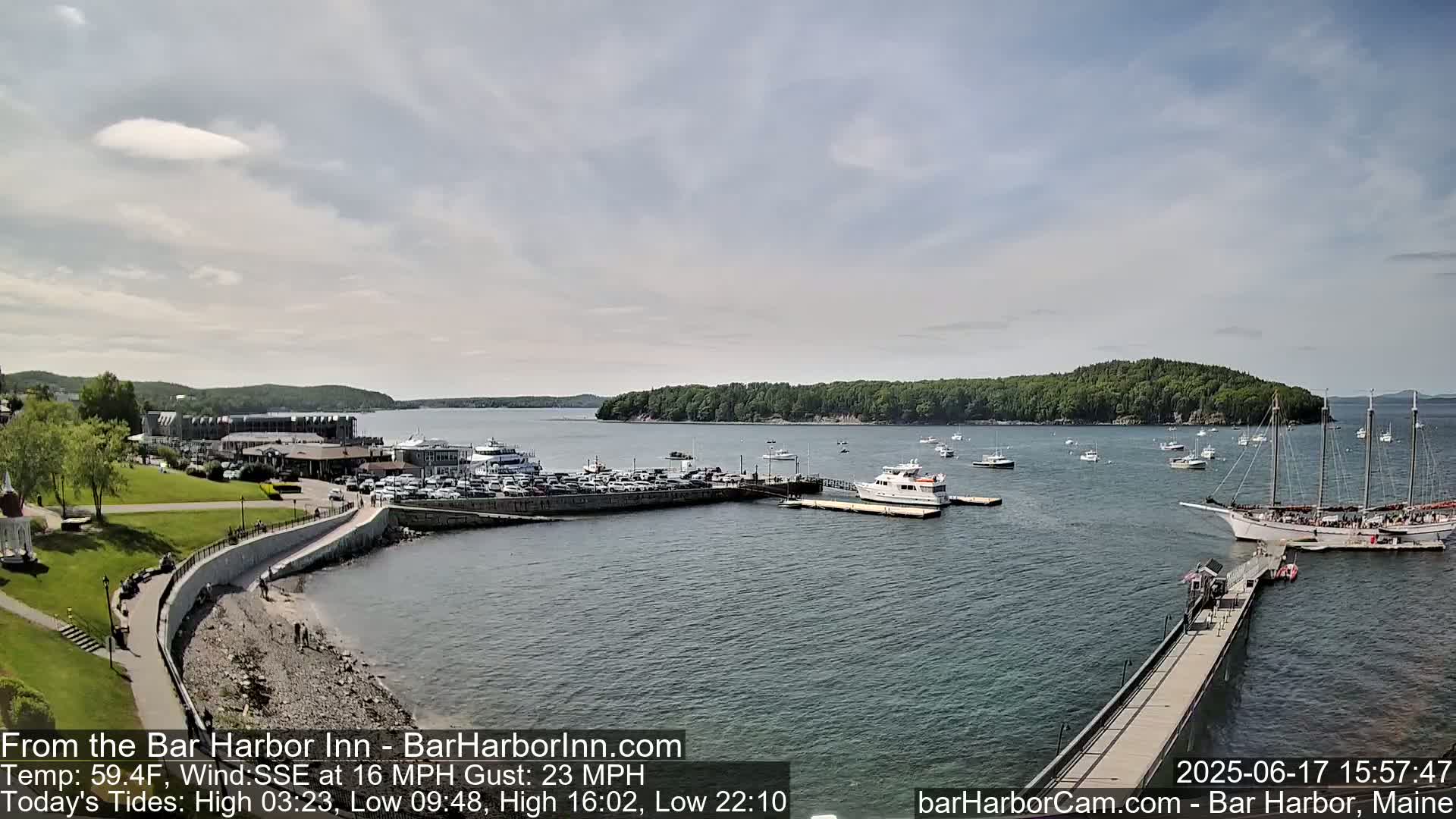 A harbor with numerous boats and a large sailboat, a parking lot, and a building is visible on a partly cloudy day with a wind blowing from the south-southeast at 16 miles per hour, gusting to 23 miles per hour.