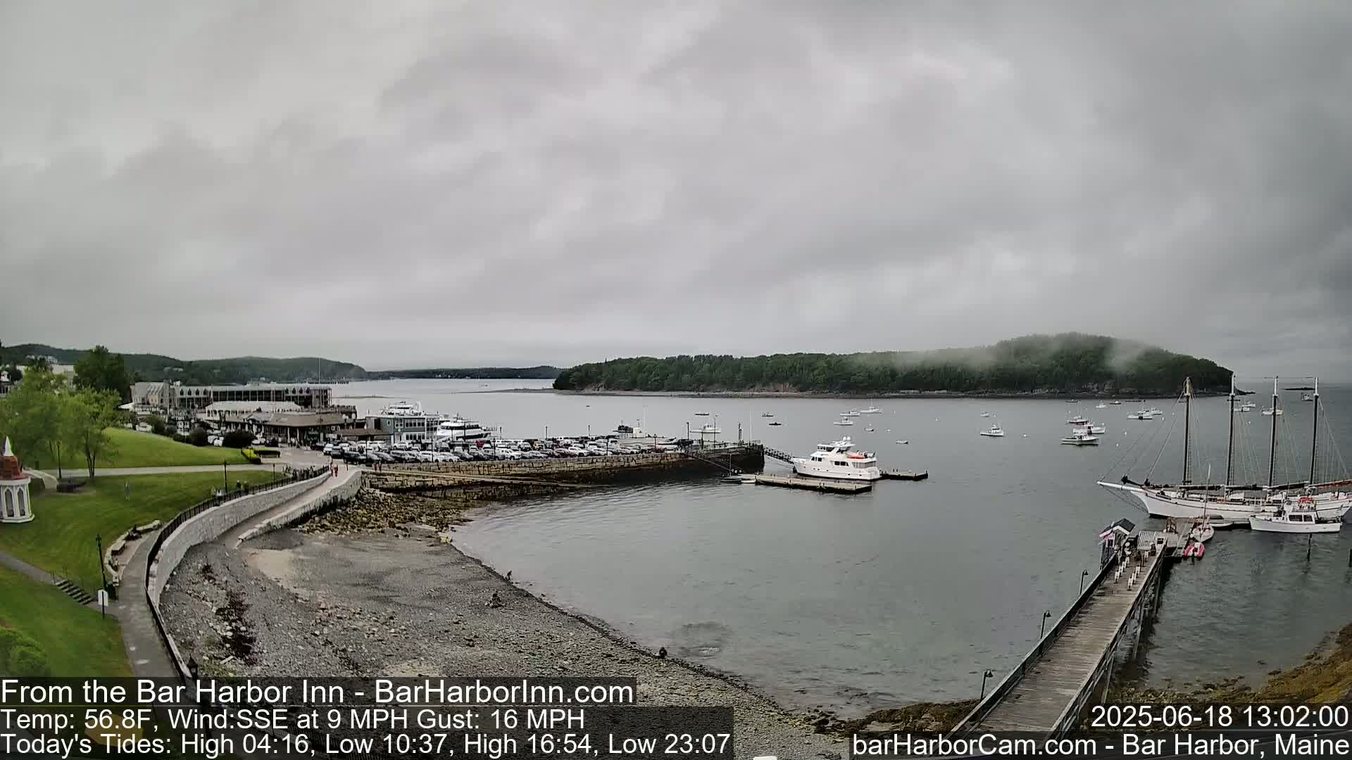 A harbor scene on an overcast day shows a coastline with buildings, a parking lot, a rocky beach, and numerous boats, including several docked sailboats and a large yacht.